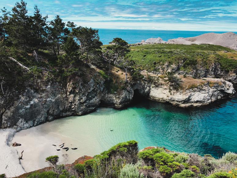 Scenic view of a rocky cove with turquoise waters, sandy beach, and lush greenery, under a blue sky.