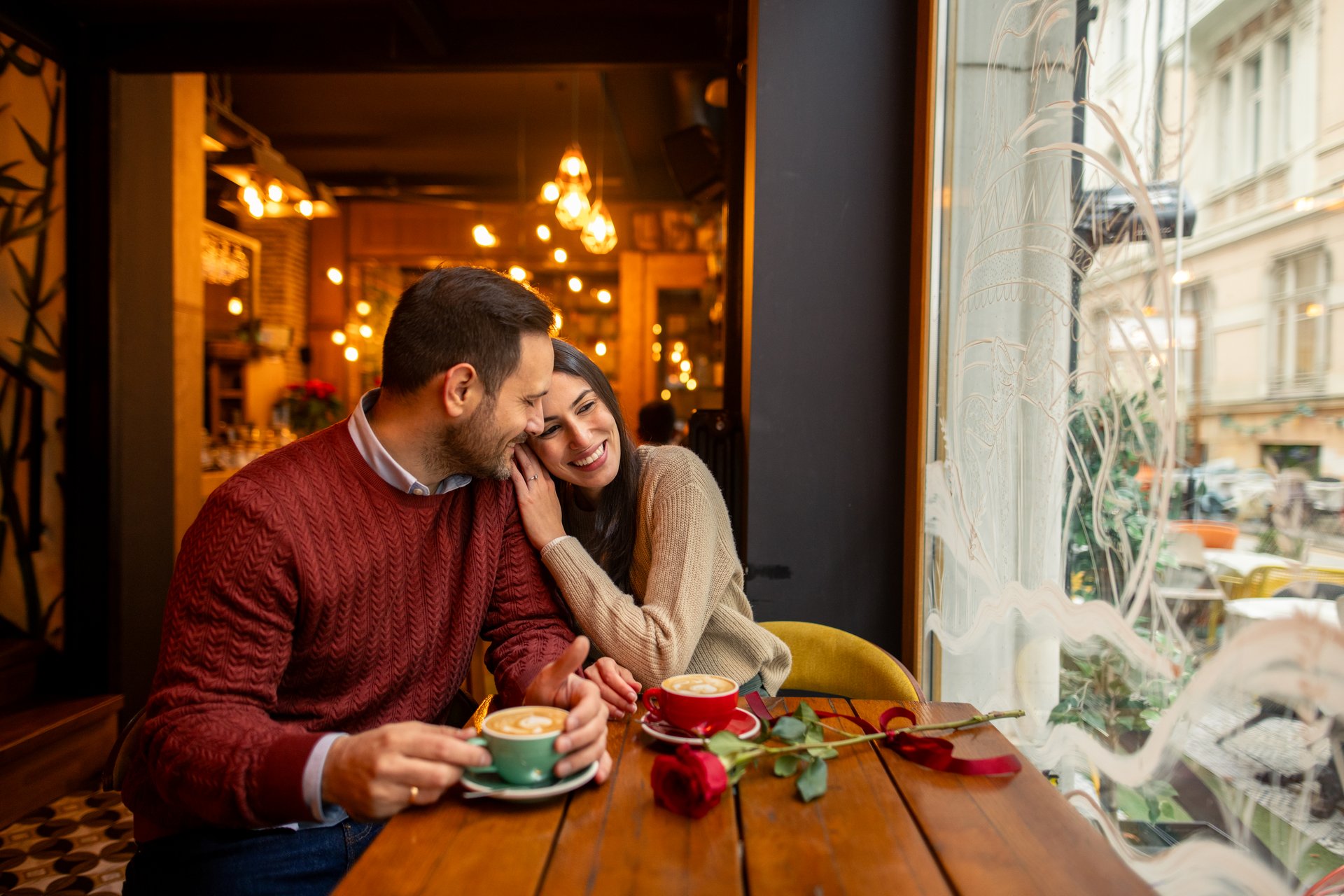 A joyful mid adult Caucasian couple, a businessman and a writer, enjoy coffee in a cozy cafe. Dressed casually with warm sweaters, they share a meaningful moment surrounded by soft lighting and romantic decor.