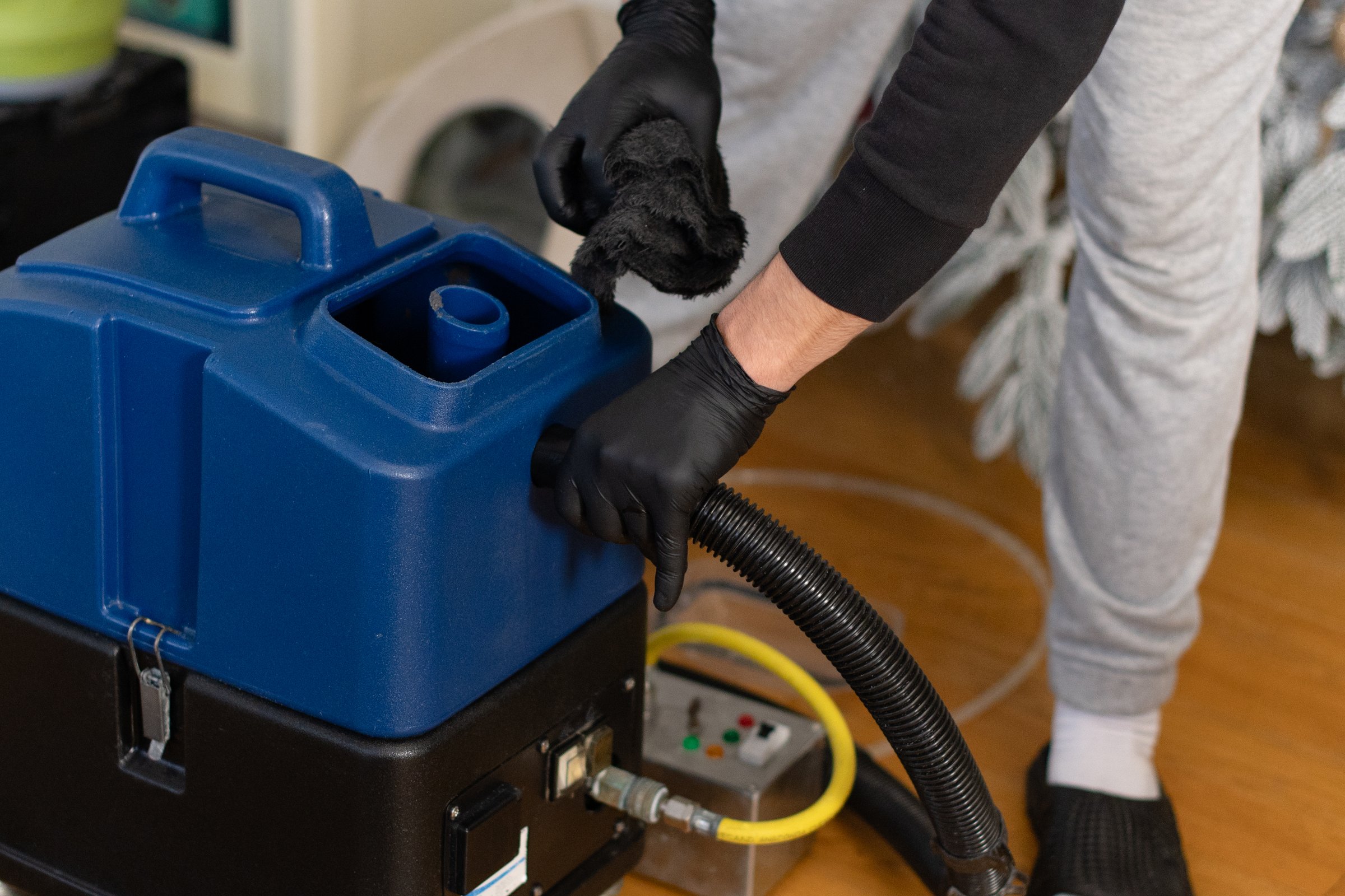 A person in gray pants and black gloves cleans a carpet using a blue machine in a home