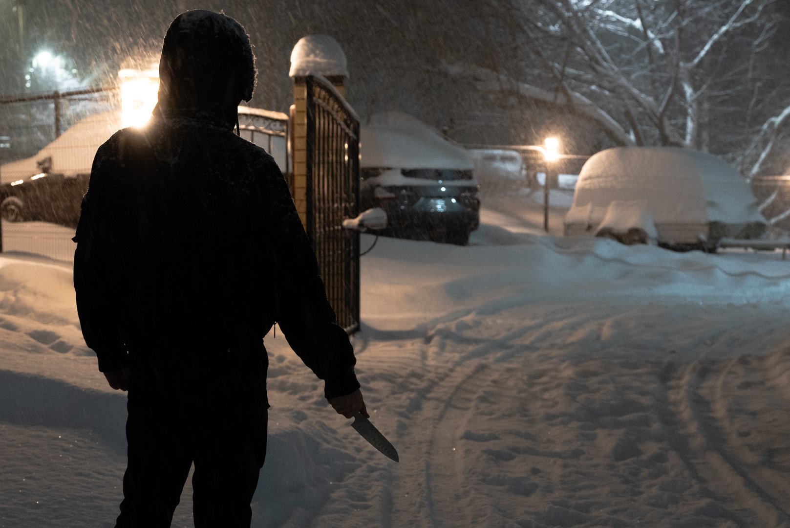 A male stalker or maniac in black clothes with a knife stands on a winter street at the gate of a parking.