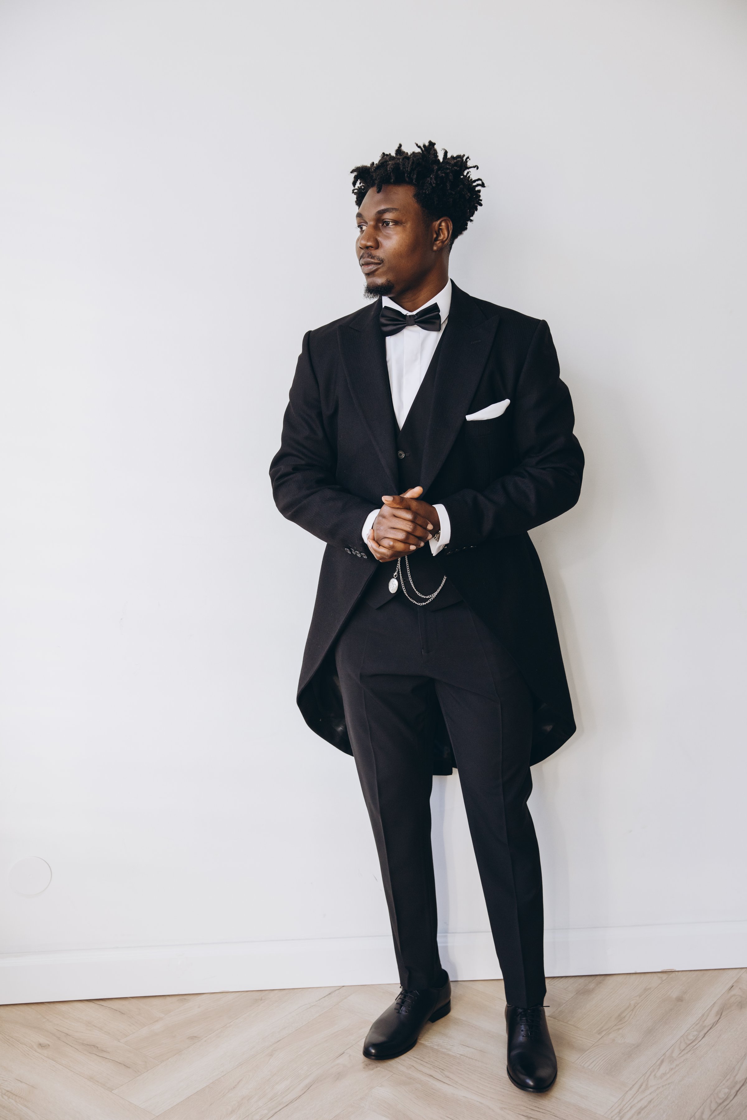 Stylish groom wearing a sophisticated black tailcoat, white shirt and bow tie, standing in a modern white room