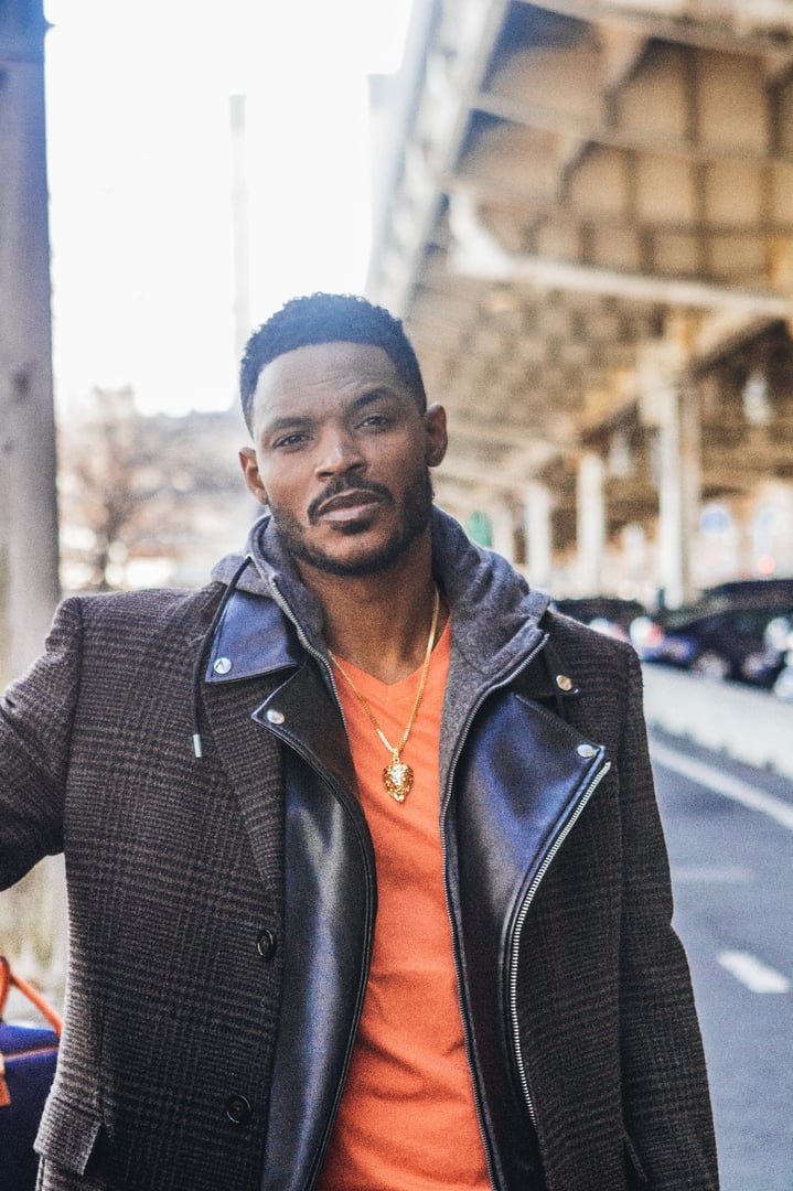 Man standing outdoors under an overpass, wearing a plaid coat, orange shirt, and a gold necklace, looking at the camera.