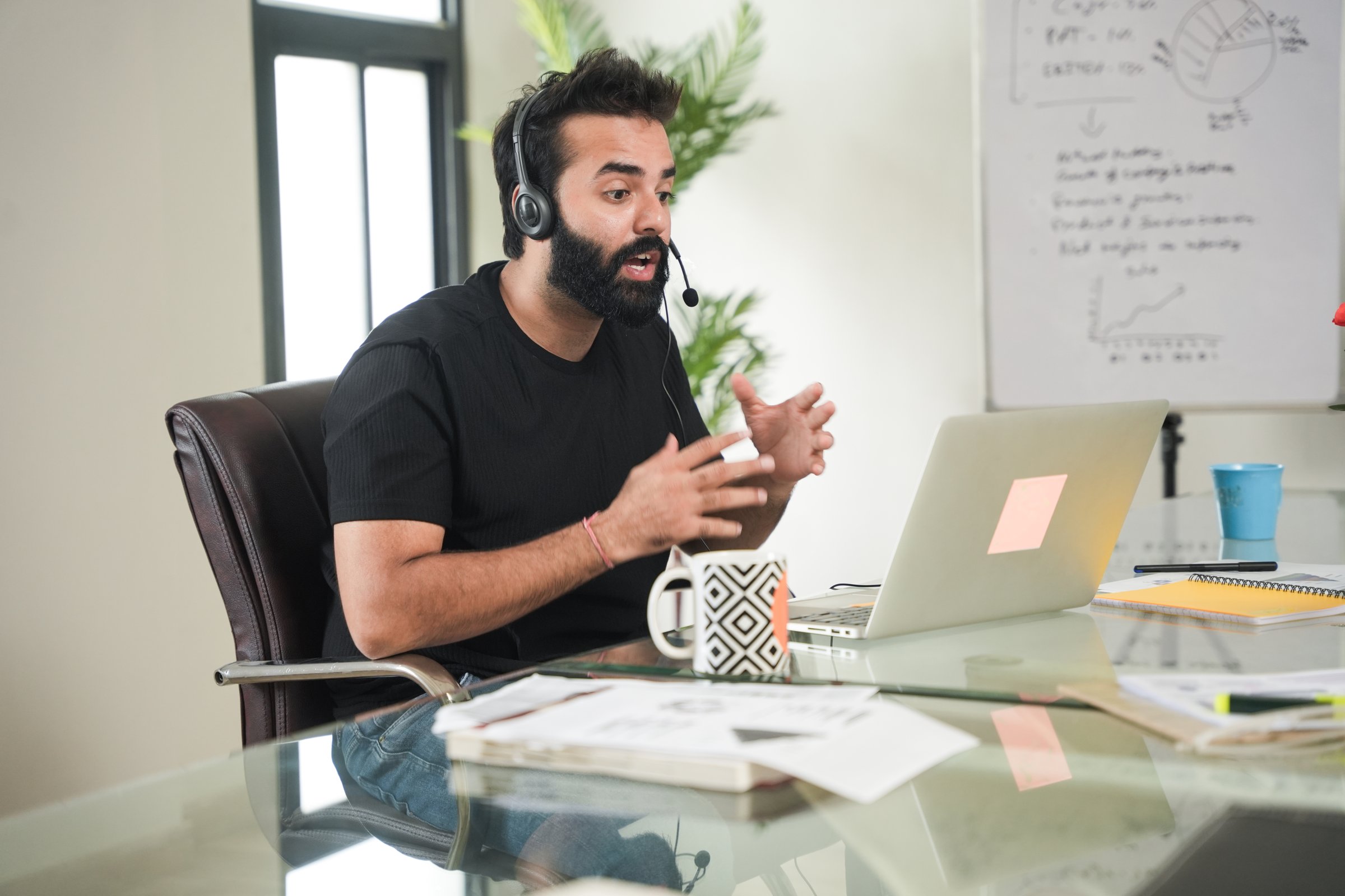 young indian man working remotely and heaving a meeting on a laptop while wearing headset. video call,  guy work at modern office or workplace, Telecommunication. Call center. Tech support services.
