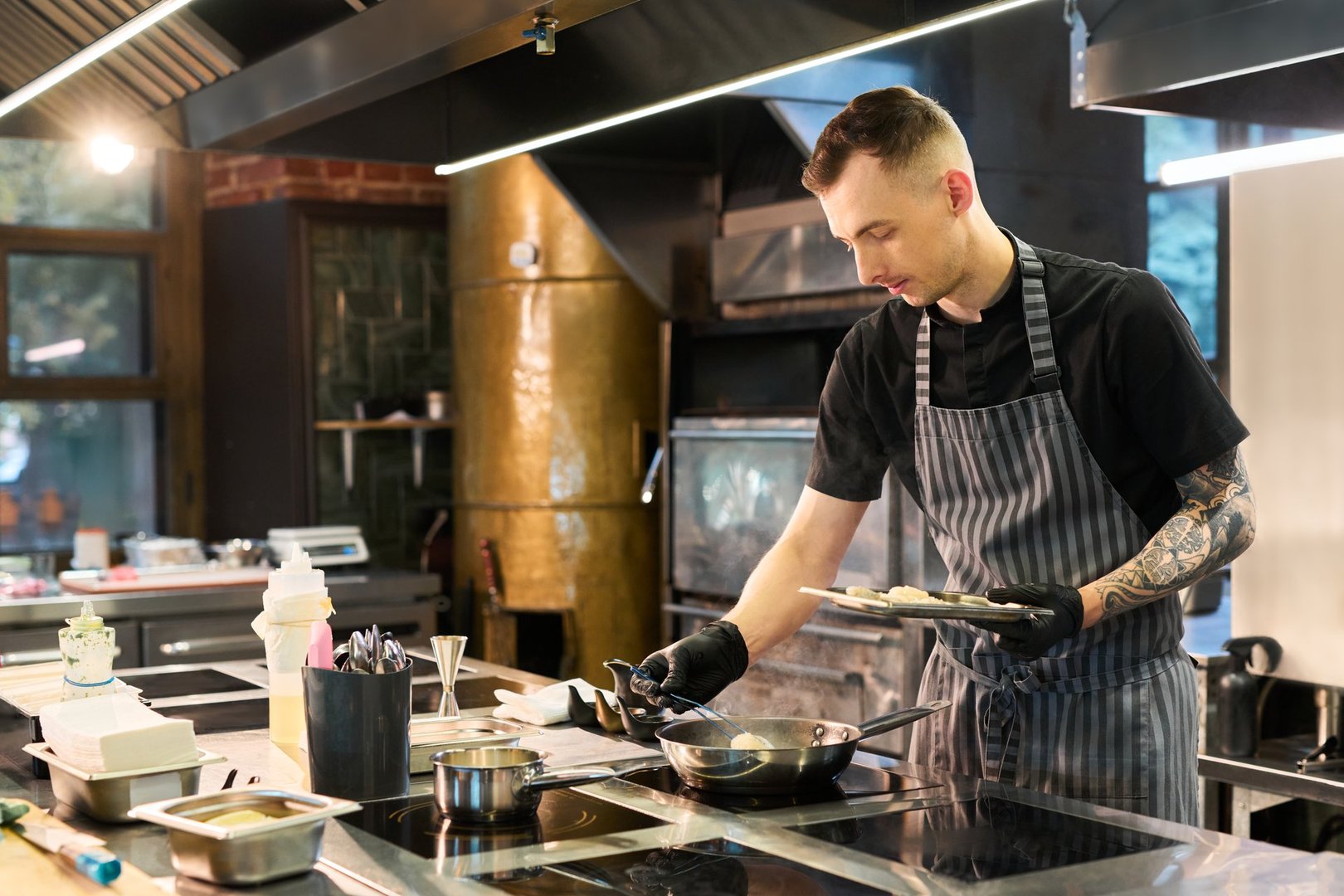 Caucasian young adult man with tattooed arm preparing food in professional kitchen, wearing apron and gloves, focused on cooking process with various utensils and ingredients visible