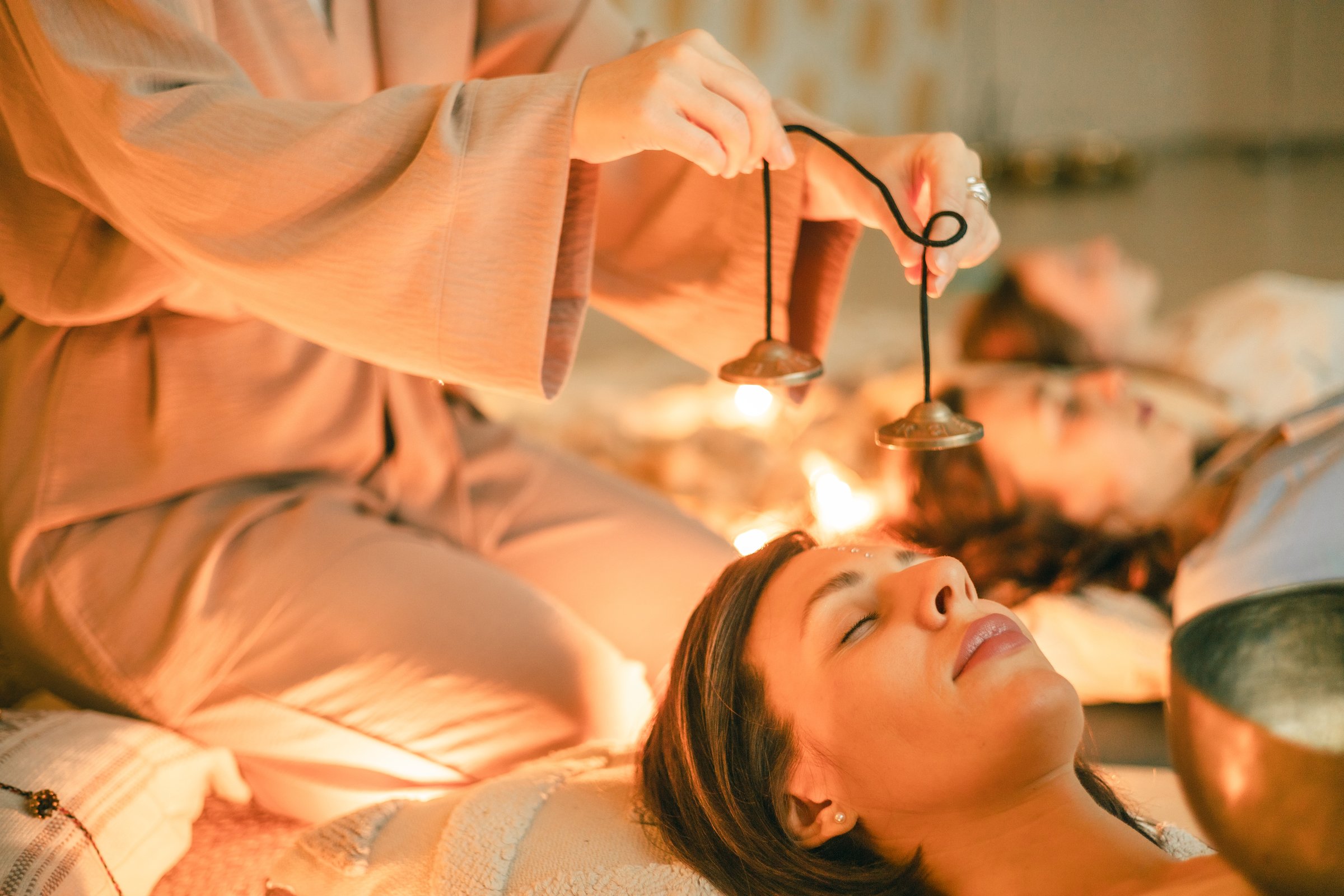 Close-up of a woman lying with eyes closed during a sound healing session as a practitioner plays tingsha bells above her, surrounded by warm lights and calming energy for deep relaxation and balance.