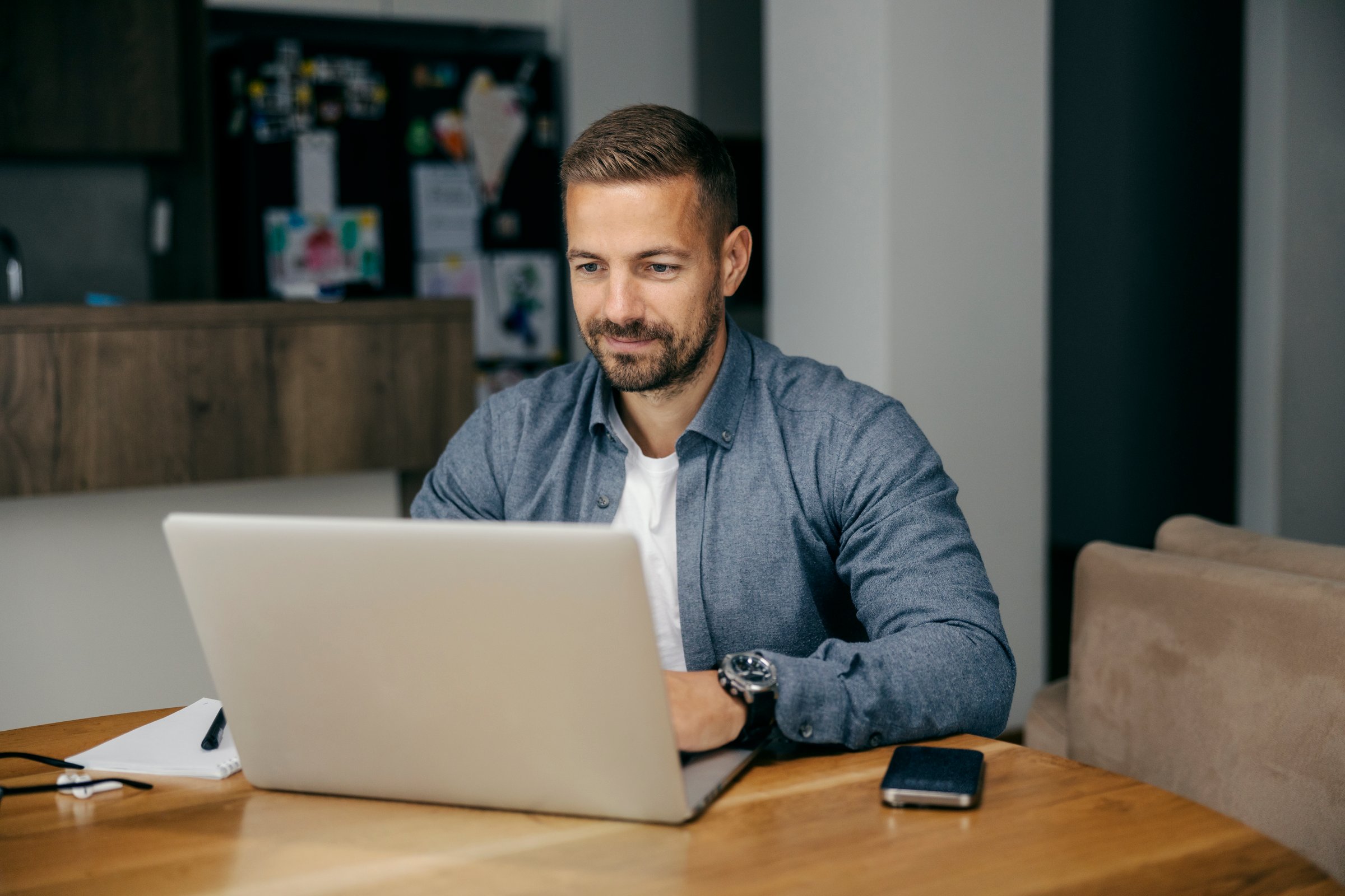 Young adult man in smart-casual shirt working from home at wooden table, focused on laptop, smiling and typing in daylight