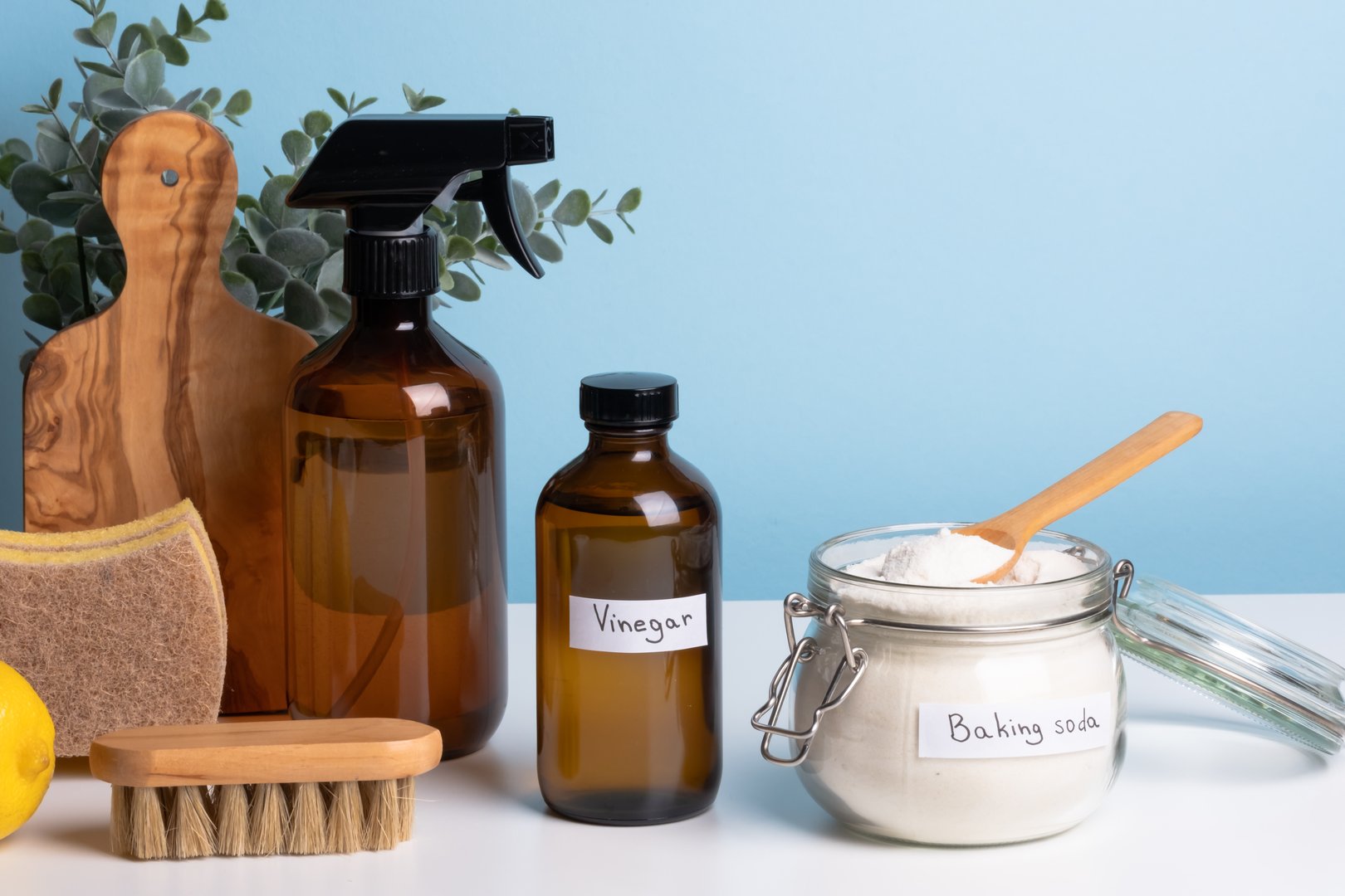A jar filled with baking soda and a wooden spoon rests on a countertop alongside a bottle of vinegar and cleaning sponges.