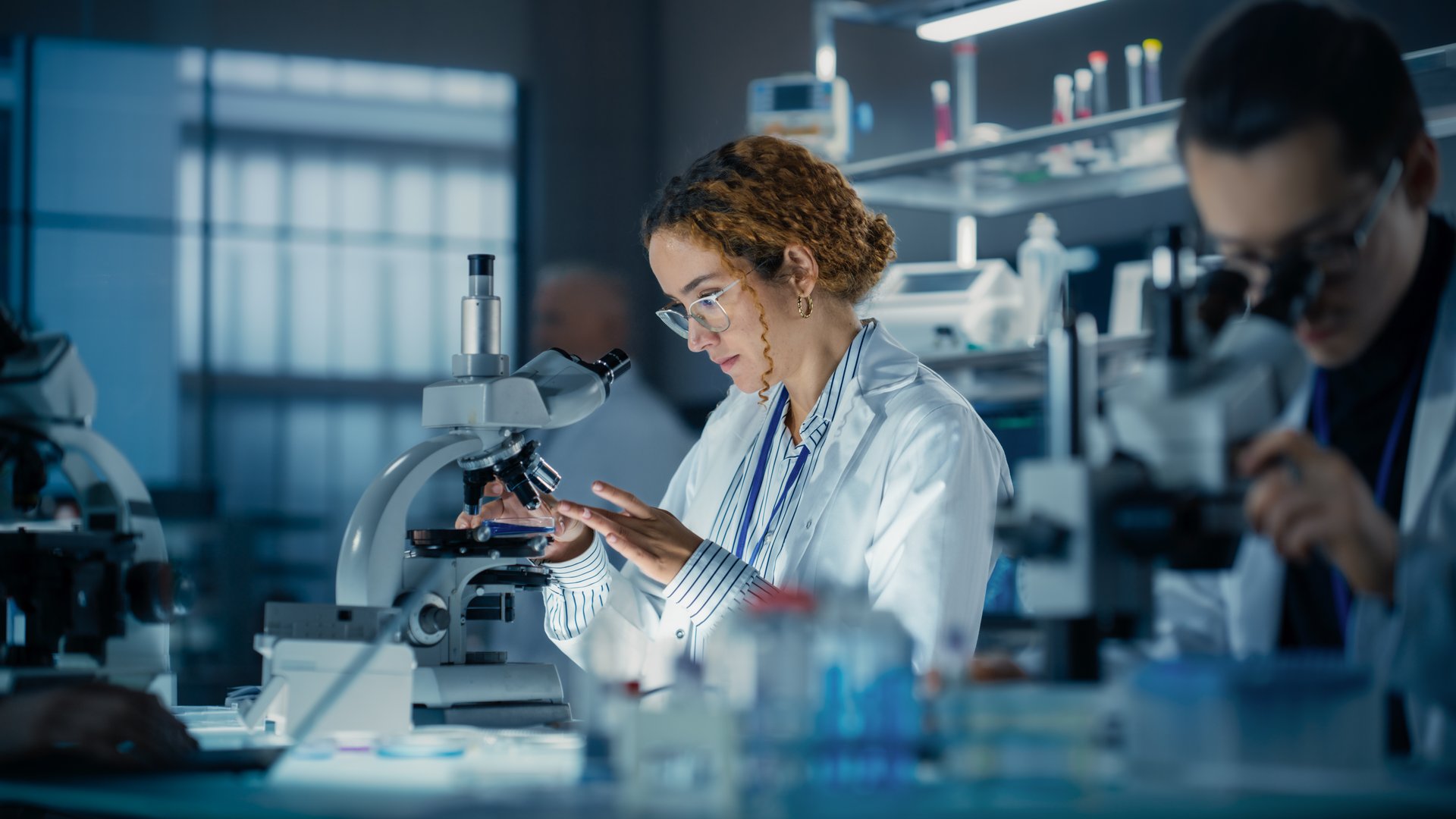 Hisplanic Female Bioengineer Looking at DNA Samples Under a Microscope in a Modern Applied Science Laboratory. Portrait of a Young Lab Engineer in White Coat Inventing New Medical Drugs
