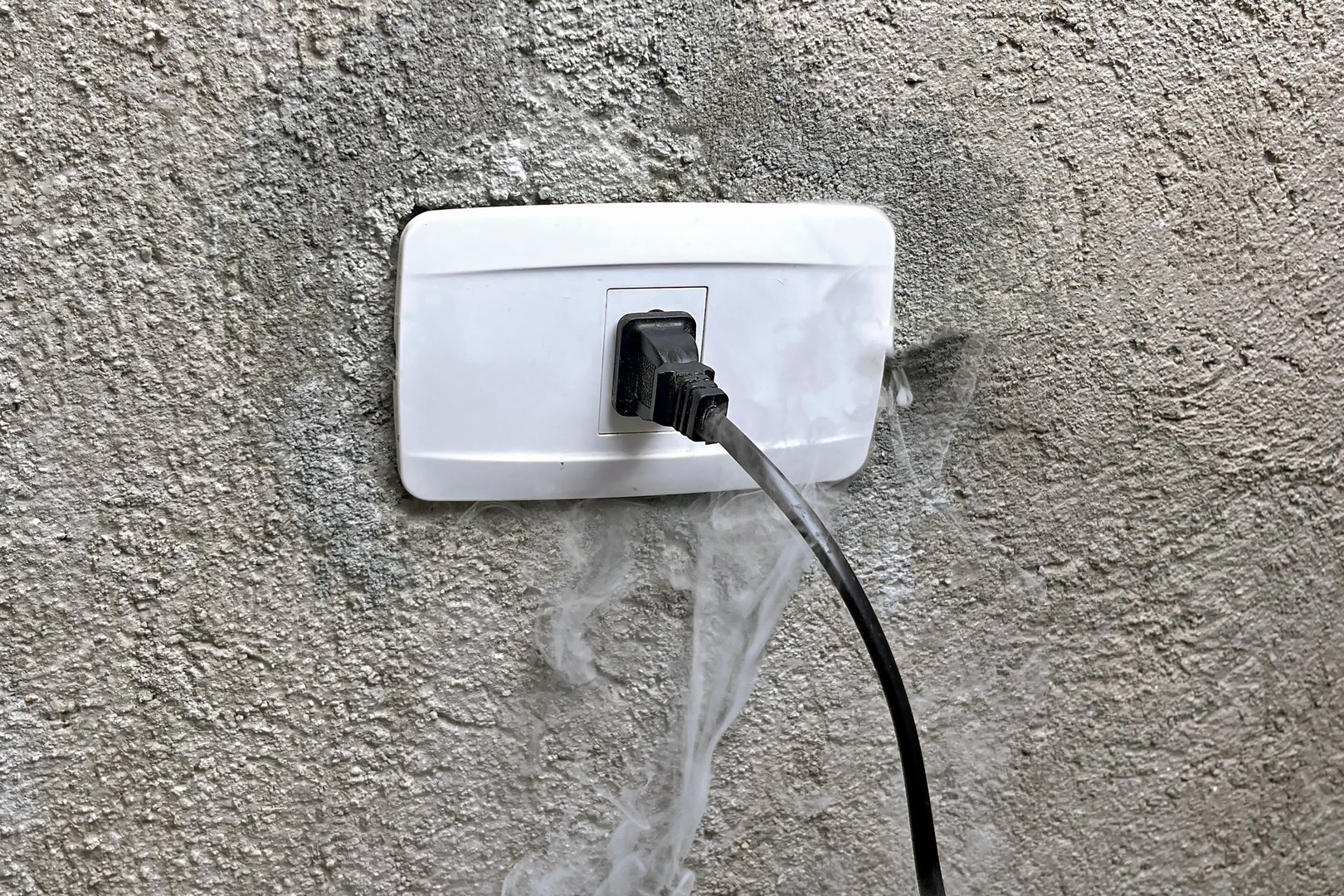 A smoking electrical outlet on a gray concrete wall from overvoltage, short circuit cause of fire.