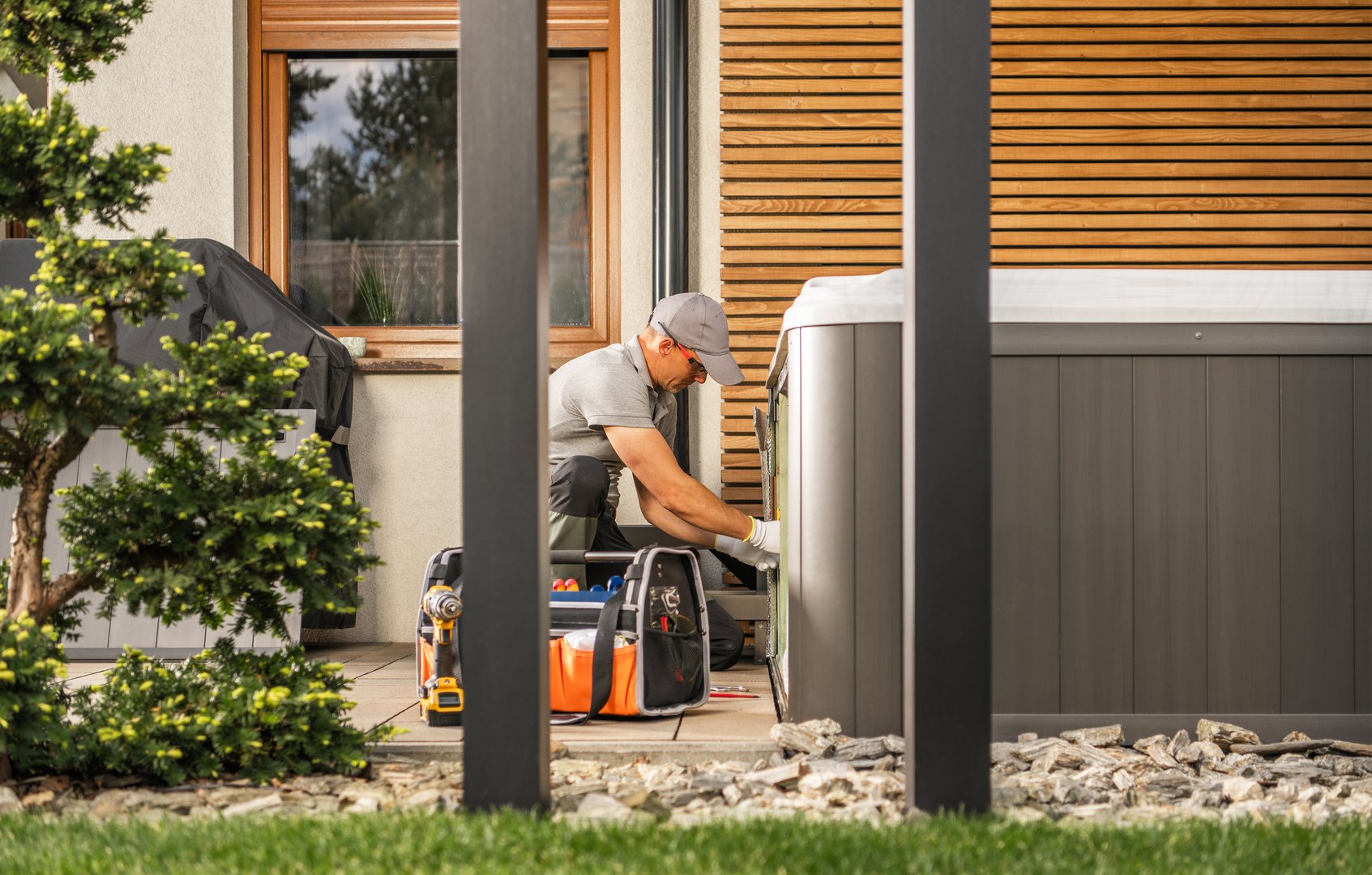 A technician works on a hot tub in a backyard setting. He is using tools to fix or maintain the hot tub. The technician is wearing a work uniform and safety glasses.