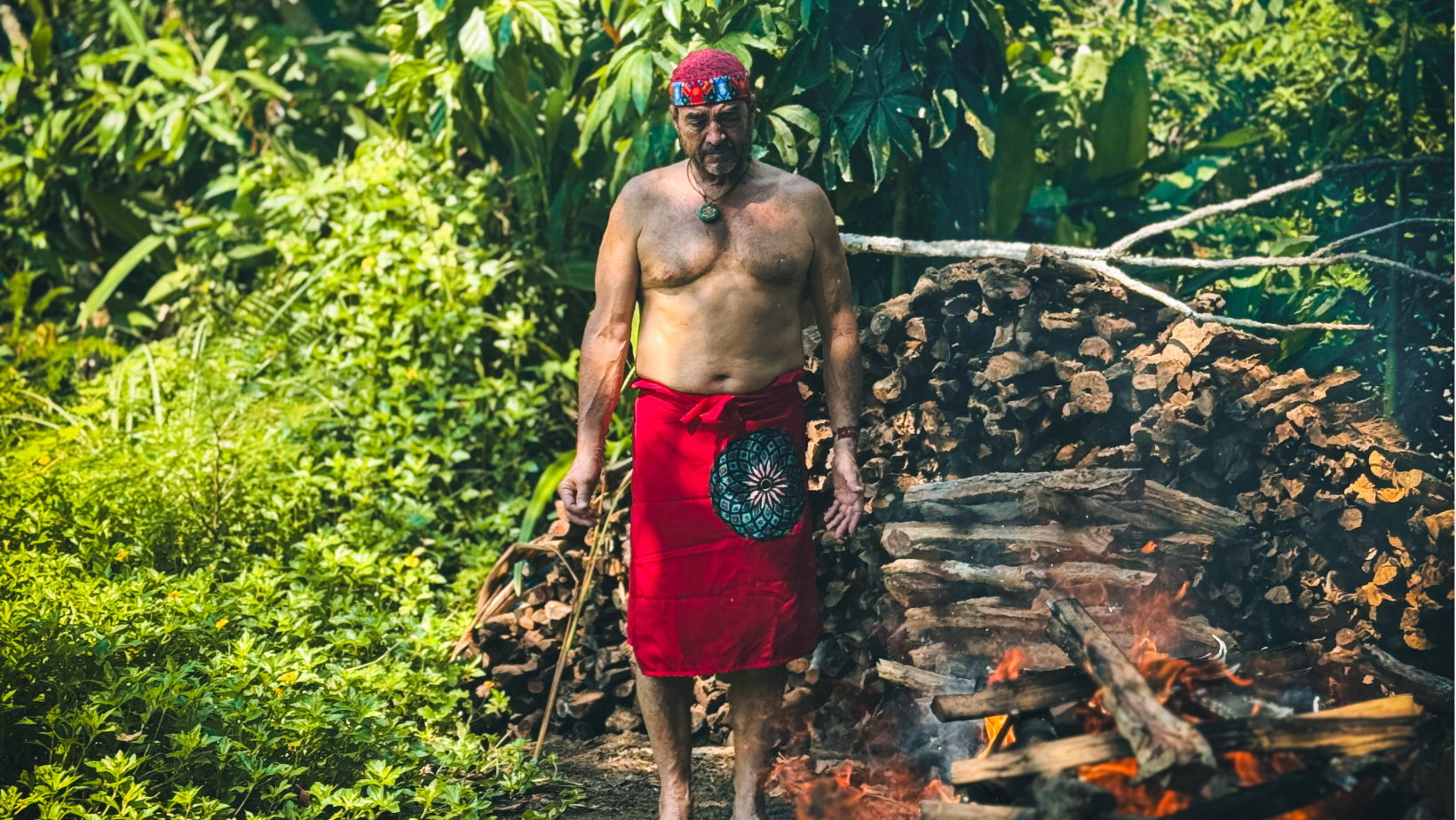 A man in traditional attire stands near a bonfire with a woodpile and lush green foliage in the background.