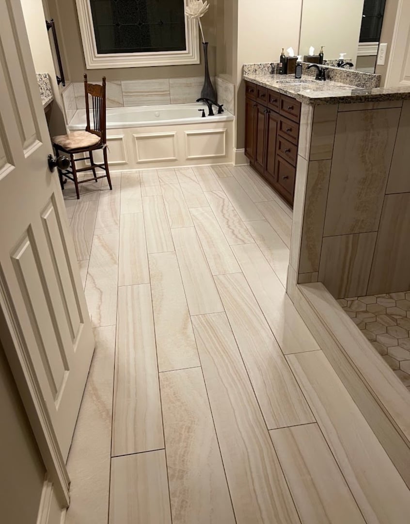 Contemporary bathroom featuring a freestanding white bathtub, double vessel sinks on a taupe vanity, and large rectangular mirror. Natural travertine-style tile covers walls and floor, complemented by louvered window shutters and sleek storage cabinet.