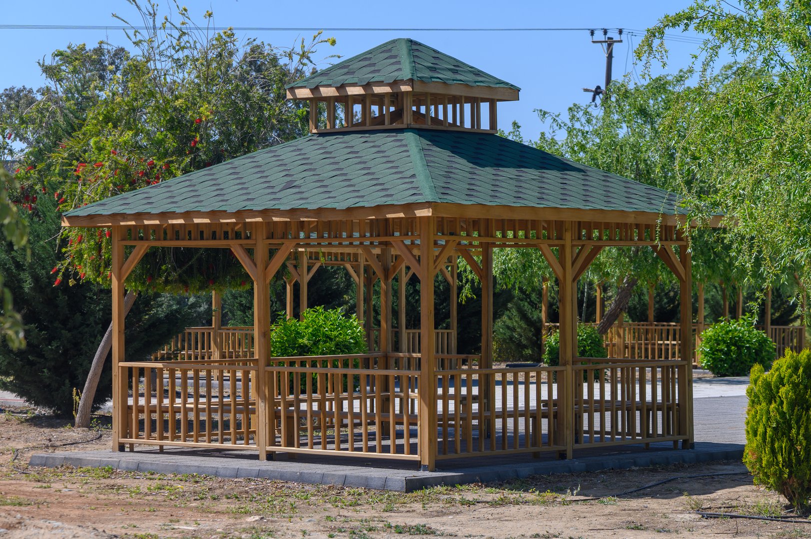 A beautifully crafted wooden gazebo stands centrally in a serene garden, surrounded by vibrant greenery and shaded by clear blue skies. It offers a perfect spot for gathering and unwinding.