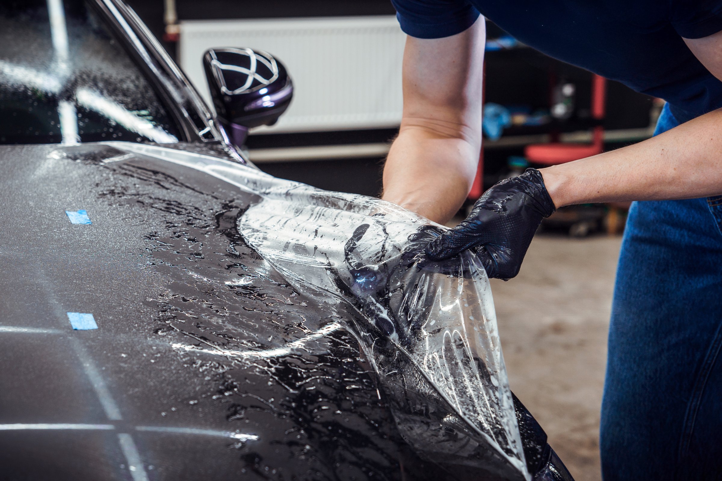 A tattooed man applies protective film to a black car at an auto detailing studio