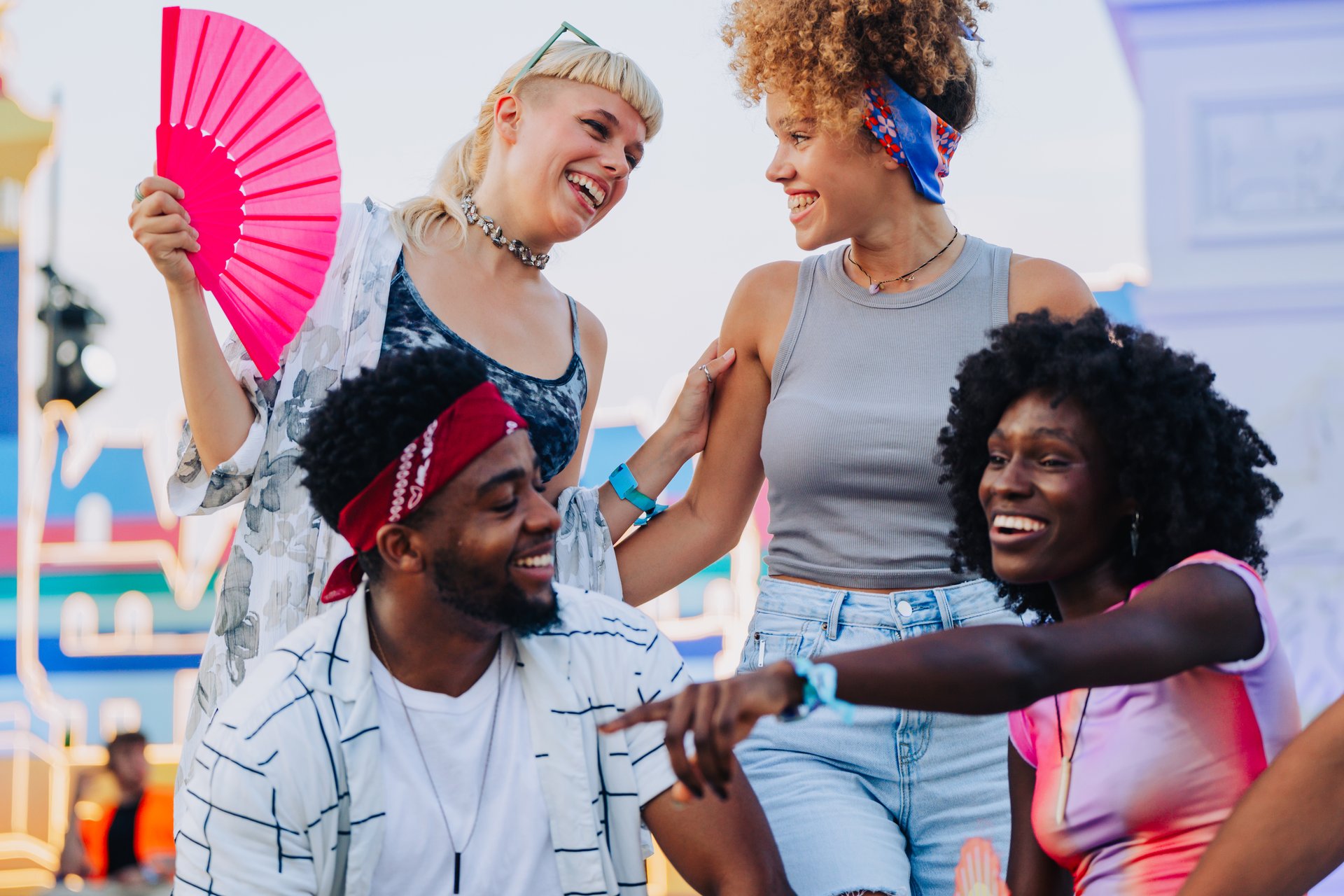 Four happy multi ethnic young adults enjoying time together at outdoor summer music festival, woman holding pink fan, colorful stage in background
