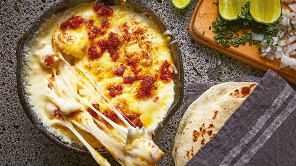 Melted cheese with chorizo in a skillet, alongside lime wedges, chopped herbs, and grilled tortillas on a cloth.