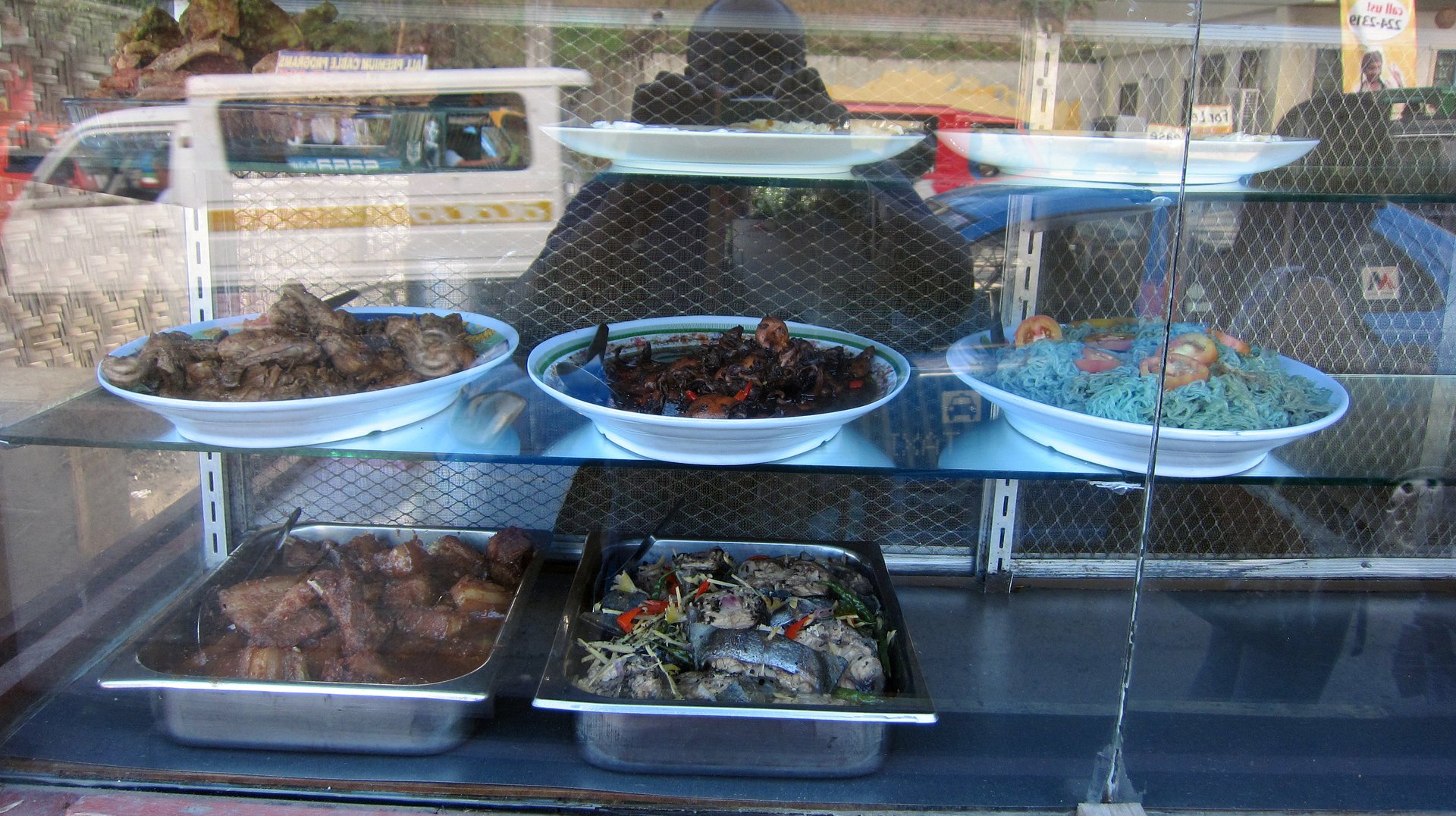 Six Filipino dishes are displayed behind a glass window at a market food stall, including braised meat, grilled fish with vegetables, and blue-colored noodles garnished with tomato slices. The vibrant arrangement evokes themes of culinary spectacle, regional texture, and tropical nourishment in a Southeast Asian street setting. Davao City, Philippines on March 31, 2012