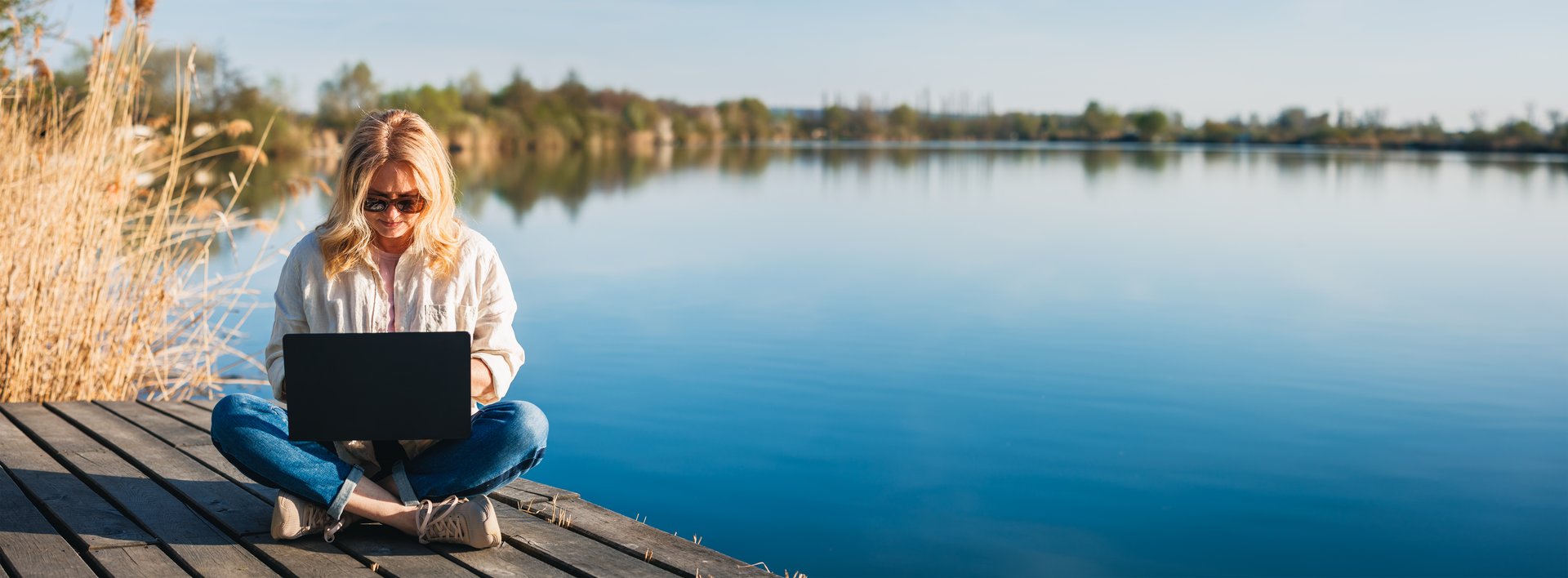 Woman working on laptop while sitting on wooden pier near water, enjoying freedom of remote job. Freelance work. Panoramic view
