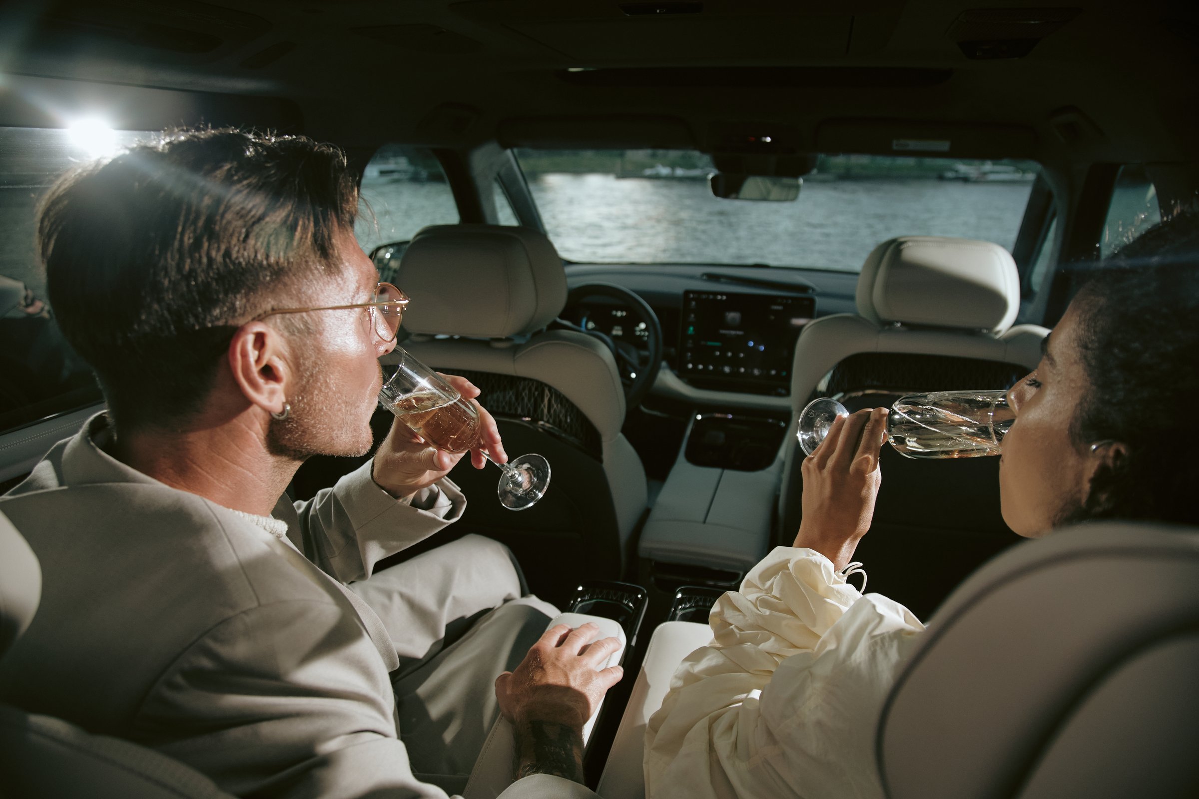 Over shoulder shot of celebrity couple enjoying sparkling champagne celebrating success sitting in back seat of car on river bank, fish eye effect