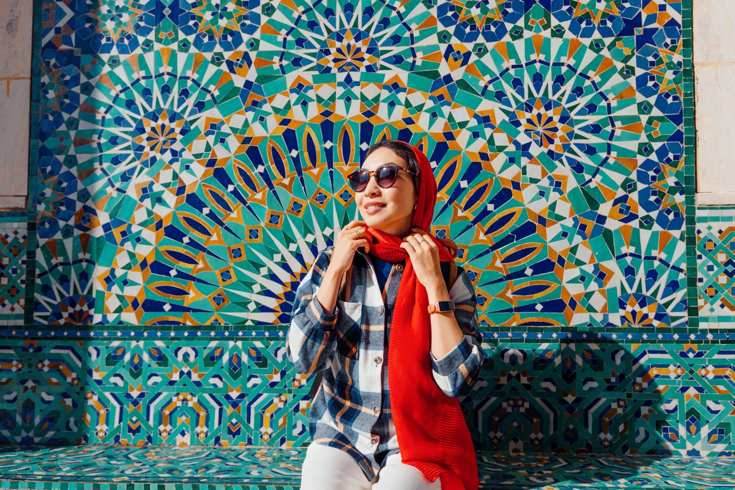 Young woman tourist wearing red scarf and sunglasses posing in front of colorful mosaic wall in Morocco