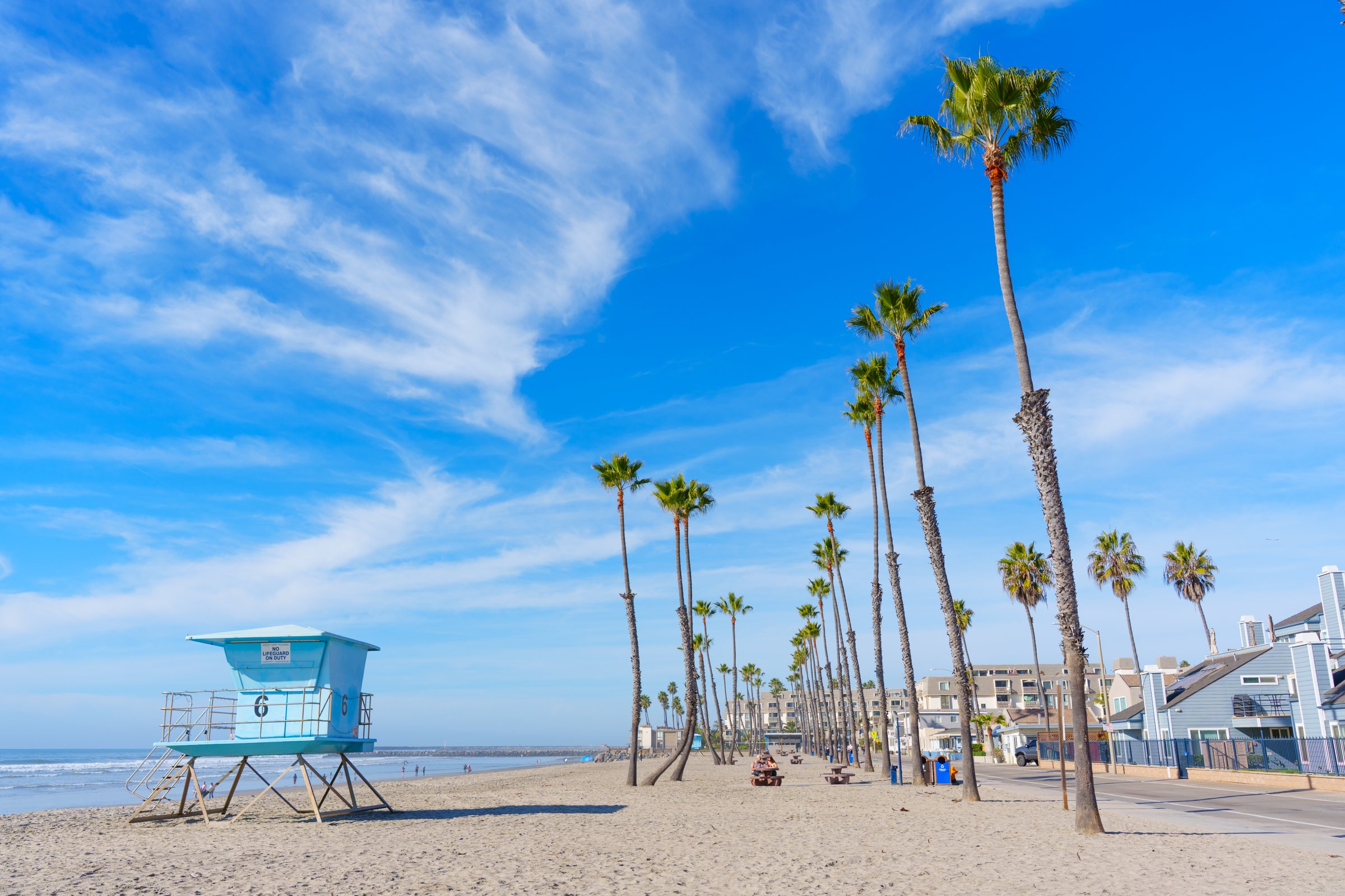 Oceanside, California - December 27, 2024: Lively beachfront in Oceanside, California showcasing lifeguard tower, palm trees, sandy shore, and bright blue sky on a sunny day.