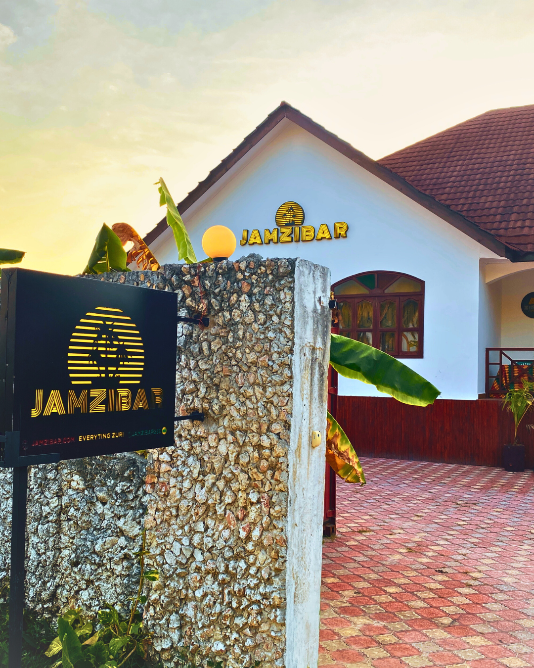 Entrance to Jamzibar with logo on wall, a white building with brown roof, and a brick-paved walkway in warm evening light.