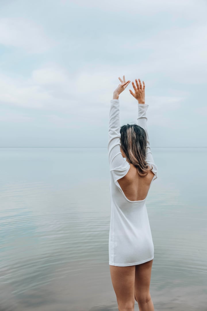 unrecognizable woman stands barefoot with hands up on wooden pier, hands up, calm sea horizon, in a white outfit, pale cloudy sky, mindfulness, wellness retreat, travel blog, vertical