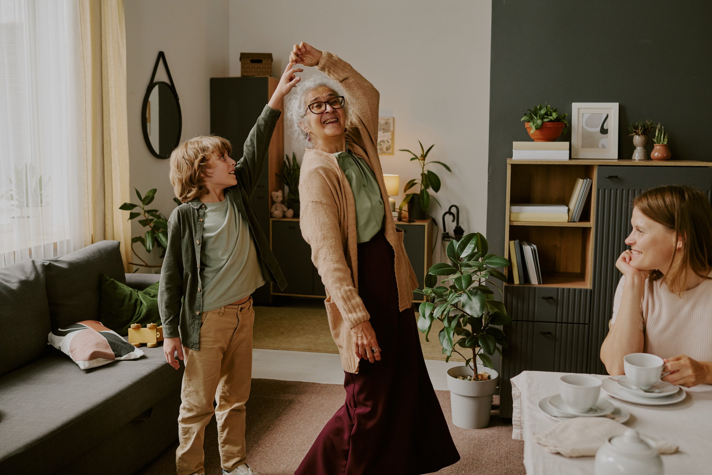 Senior Caucasian woman dancing with young Caucasian boy in living room while middle aged Caucasian woman sitting at table watching them smiling, family spending time together indoors