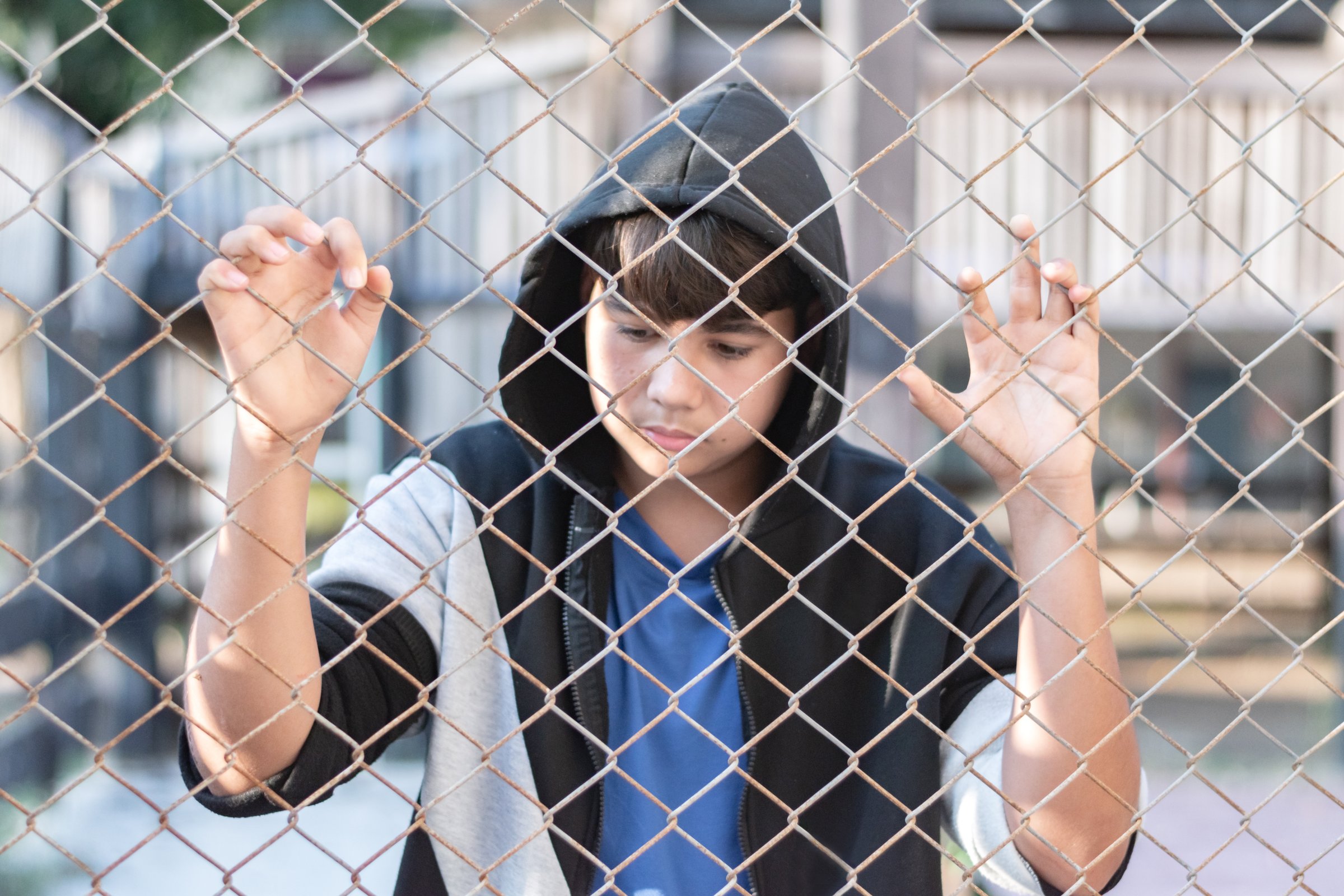 A young boy wearing a gray hoody stands with his hands on a metal fence in a local detention center, looking sad and wanting his freedom.