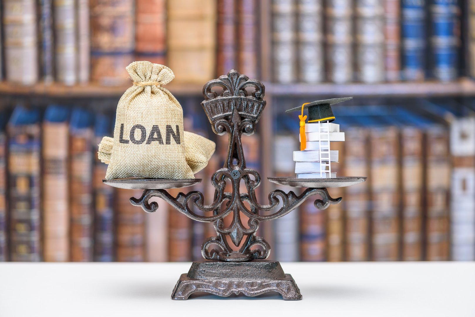 Student loan, education costs, financial burden related to higher education : A balance scale with a sack labeled 'LOAN' on one side and a stack of books topped with a graduation cap on the other.