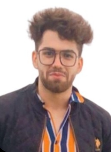 A man with curly hair, beard, and glasses wearing a black jacket over a striped shirt, against a white background.