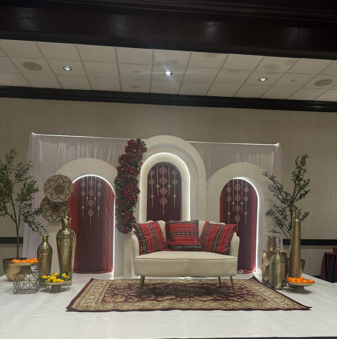 Ornate wedding stage setup with a white sofa, red pillows, decorative vases, and floral arches, flanked by artificial trees.