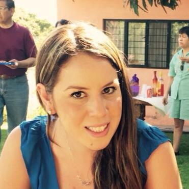 Woman with long brown hair smiling outdoors at a gathering, with people and a table with decorations in the background.