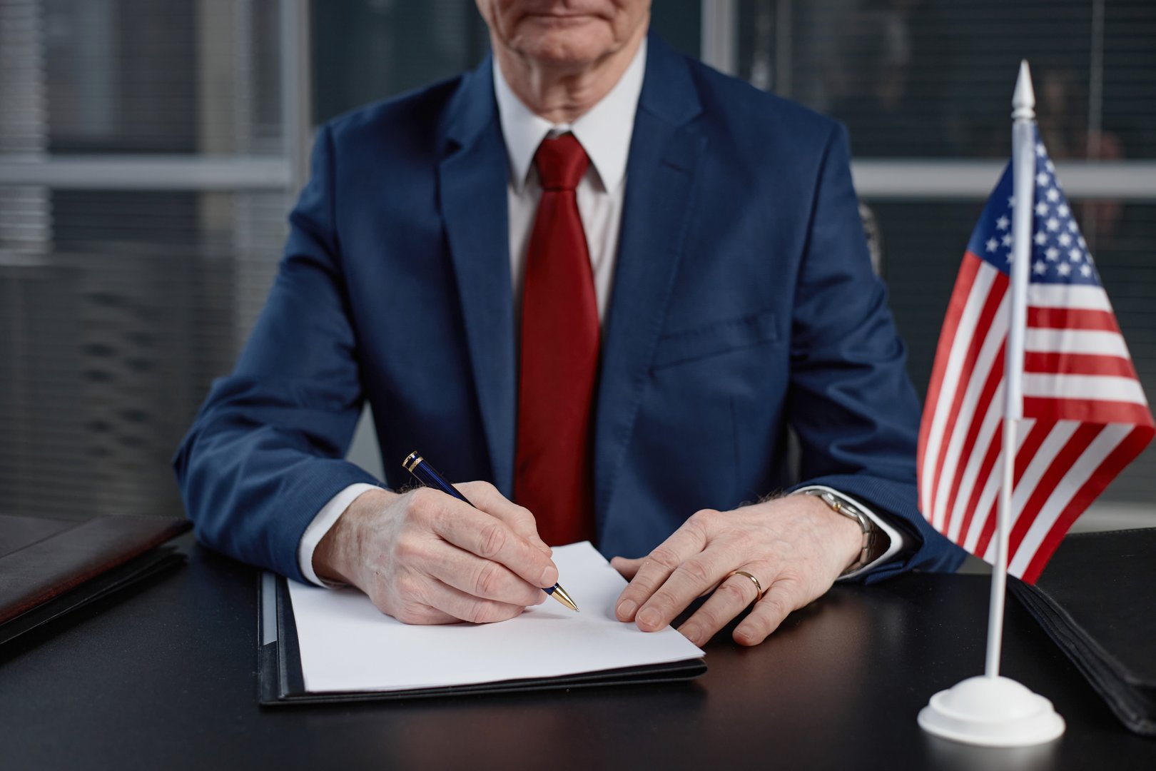 Cropped shot of Caucasian senior male politician wearing suit signing document with pen sitting at desk with small American flag at office