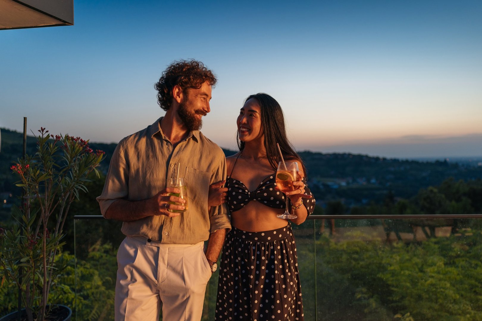 Happy couple is standing on a balcony with drinks, looking at each other and enjoying the sunset over the landscape