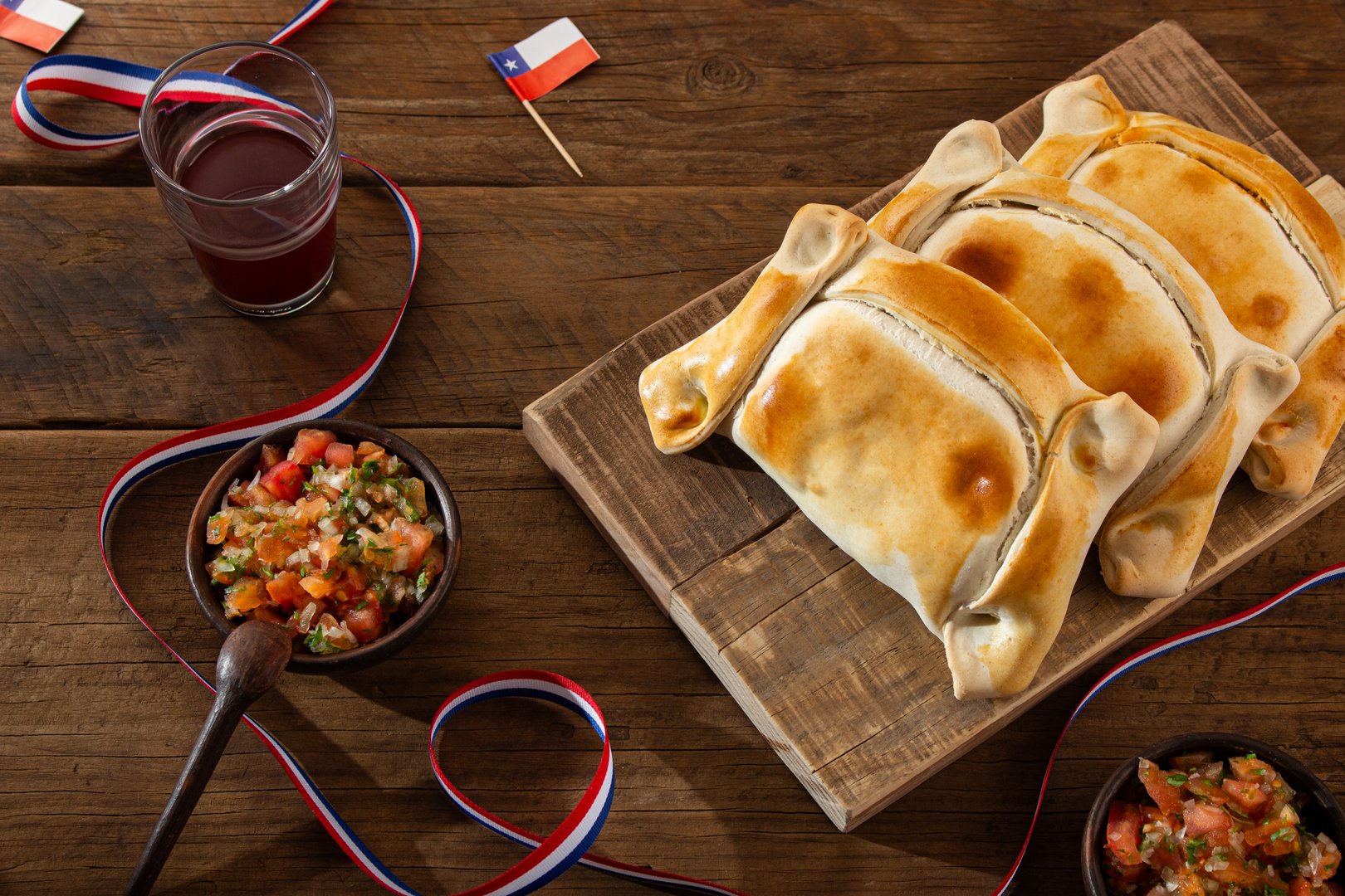 Traditional Chilean empanadas with pebre sauce and a drink presented on a rustic wooden table. Chilean national holidays, September 18th.