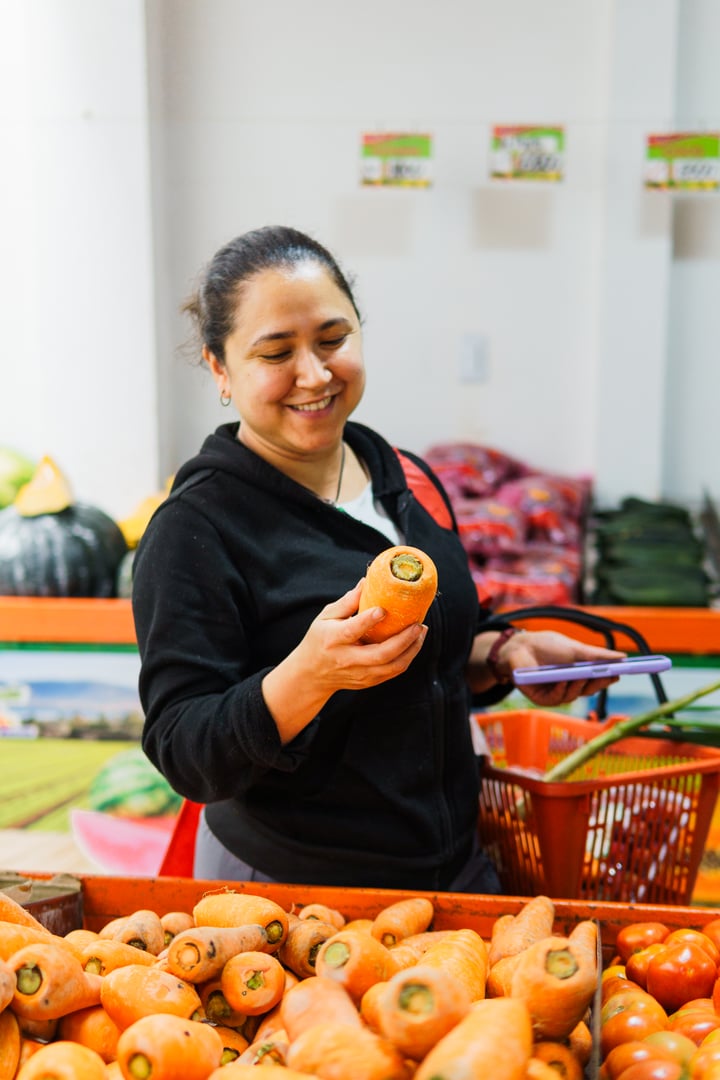 Portrait of a domestic worker choosing fruits and vegetables at the market. The woman is looking at a carrot and is smiling. He has a market basket in his arm.