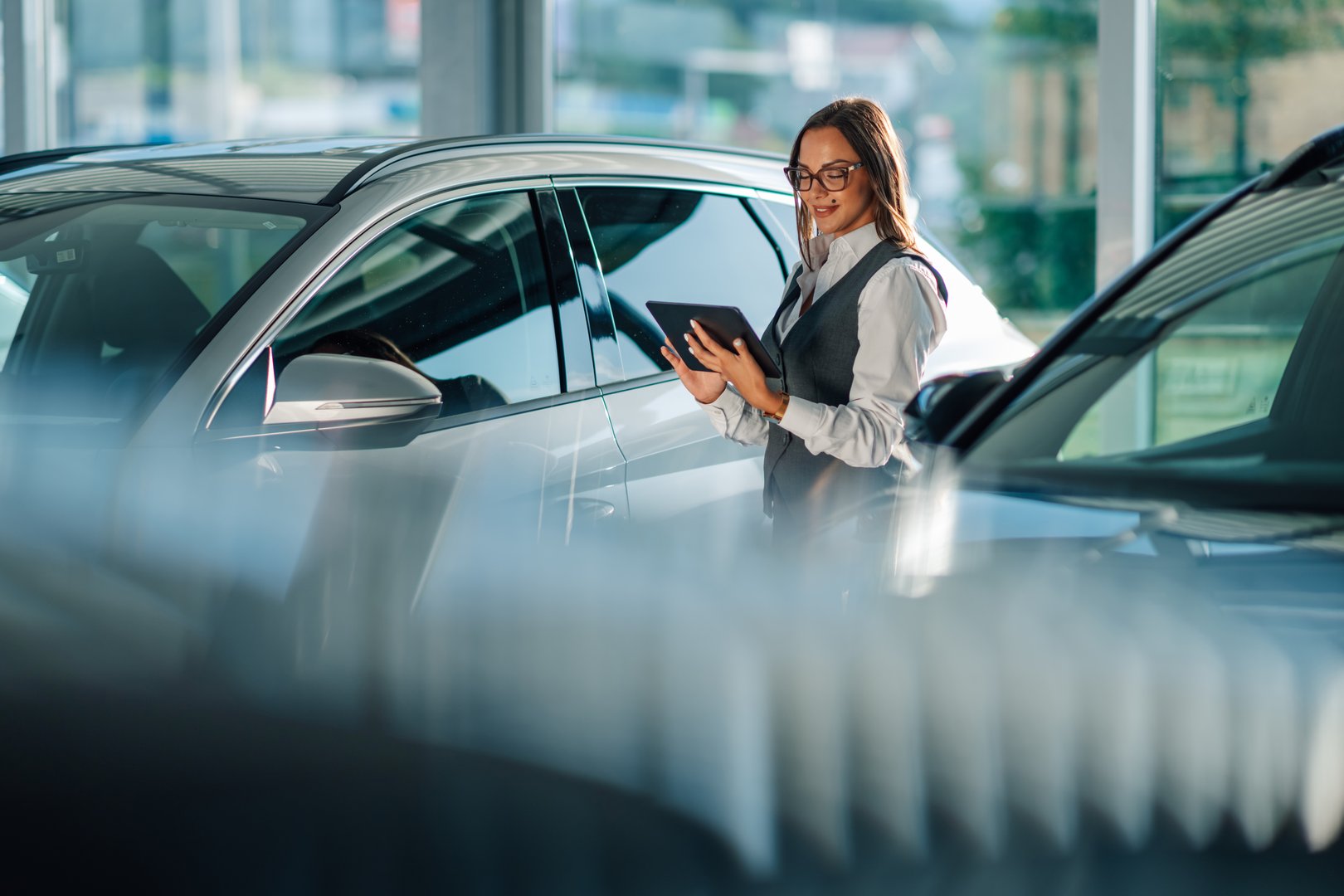 A professional woman is seen utilizing a tablet to assess cars in a modern dealership showroom. The atmosphere conveys innovation and precision in the art of automotive evaluation and sales.