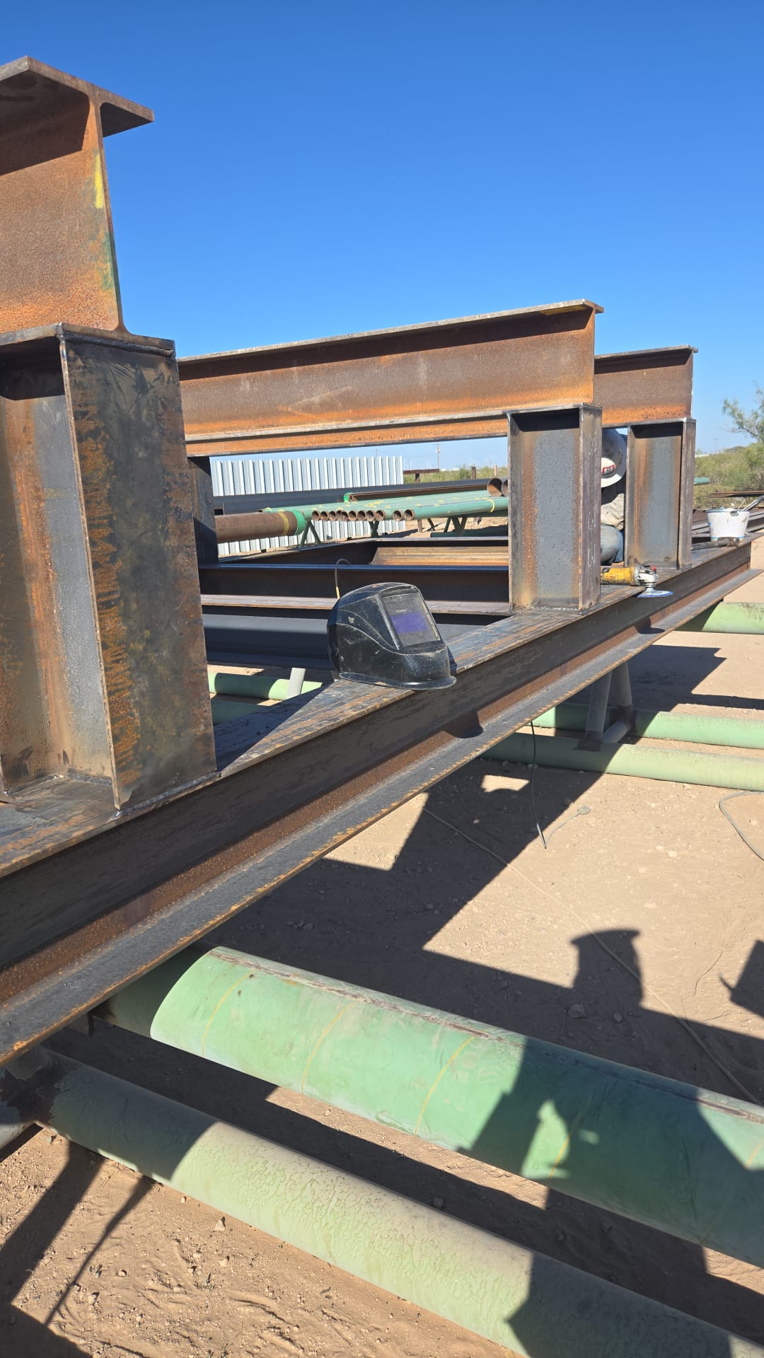 Metal beams on a construction site with a welding helmet resting on one beam. Blue sky and some vegetation in the background.