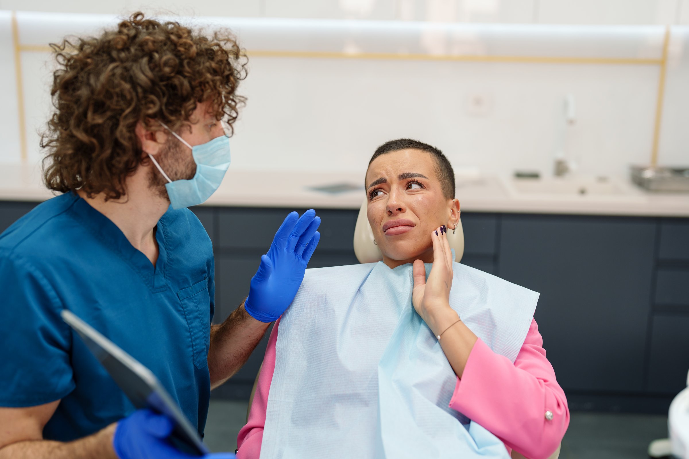 A Caucasian male dentist engages with a Caucasian female patient in a modern clinic setting. They discuss treatment options using a digital tablet. The patient wears a pink jacket and protective bib, while the dentist dons blue scrubs and a mask.