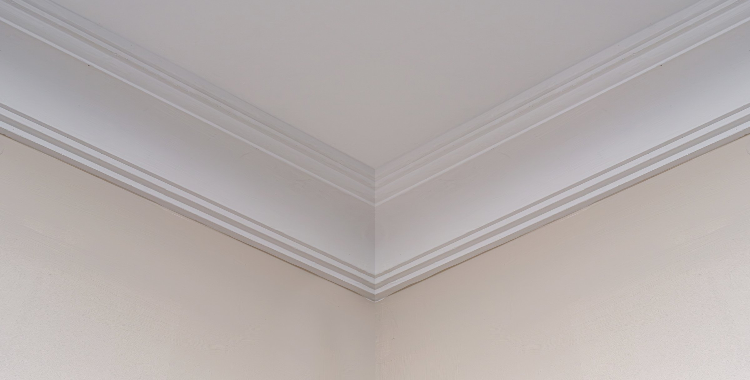 Interior corner of a room showing detailed white crown molding against beige painted walls. The trim highlights clean design, symmetry and elegant finishing in home or office interiors.