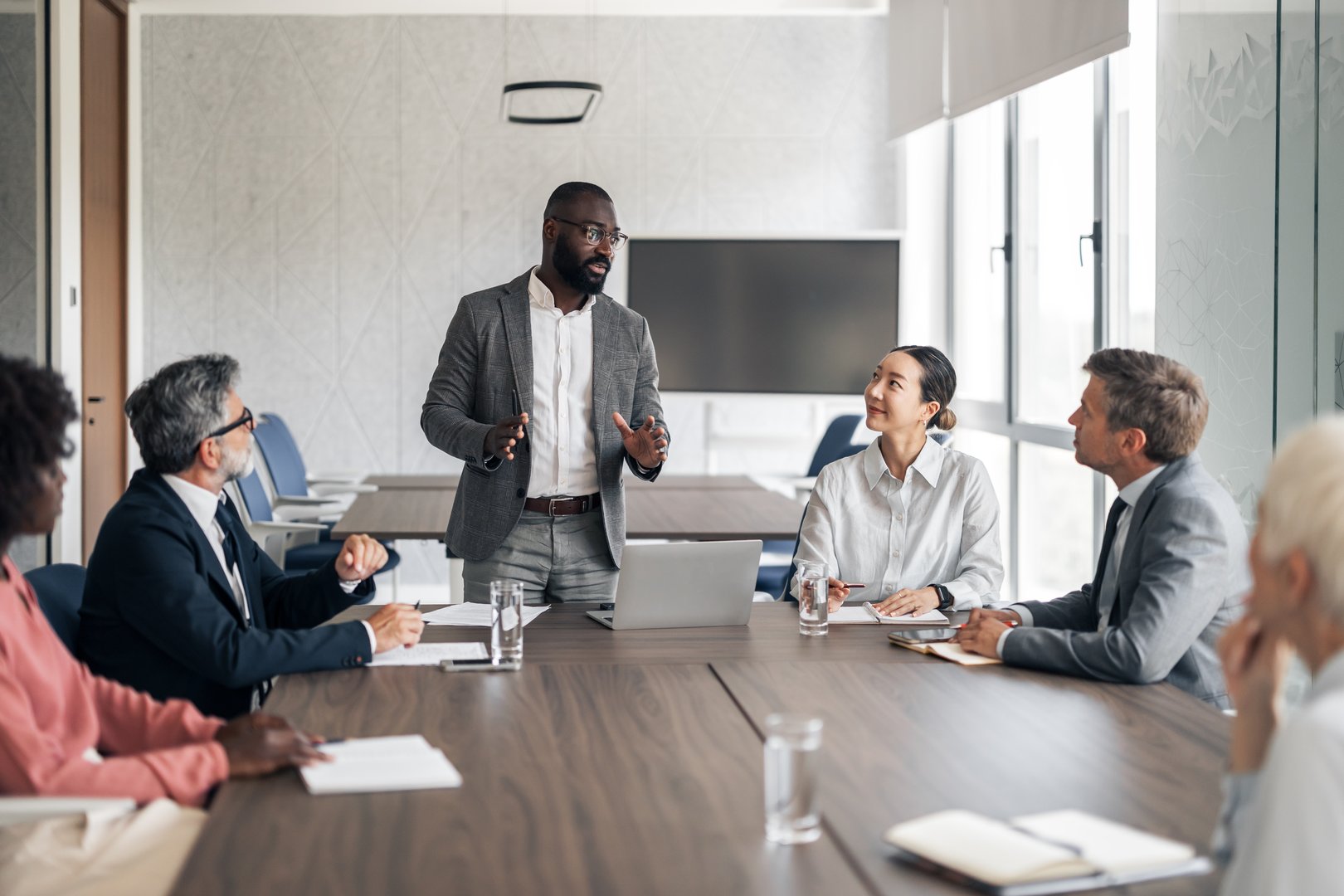 Black businessman presenting strategy to a diverse team in a modern boardroom, colleagues listening attentively around a conference table with laptop and documents