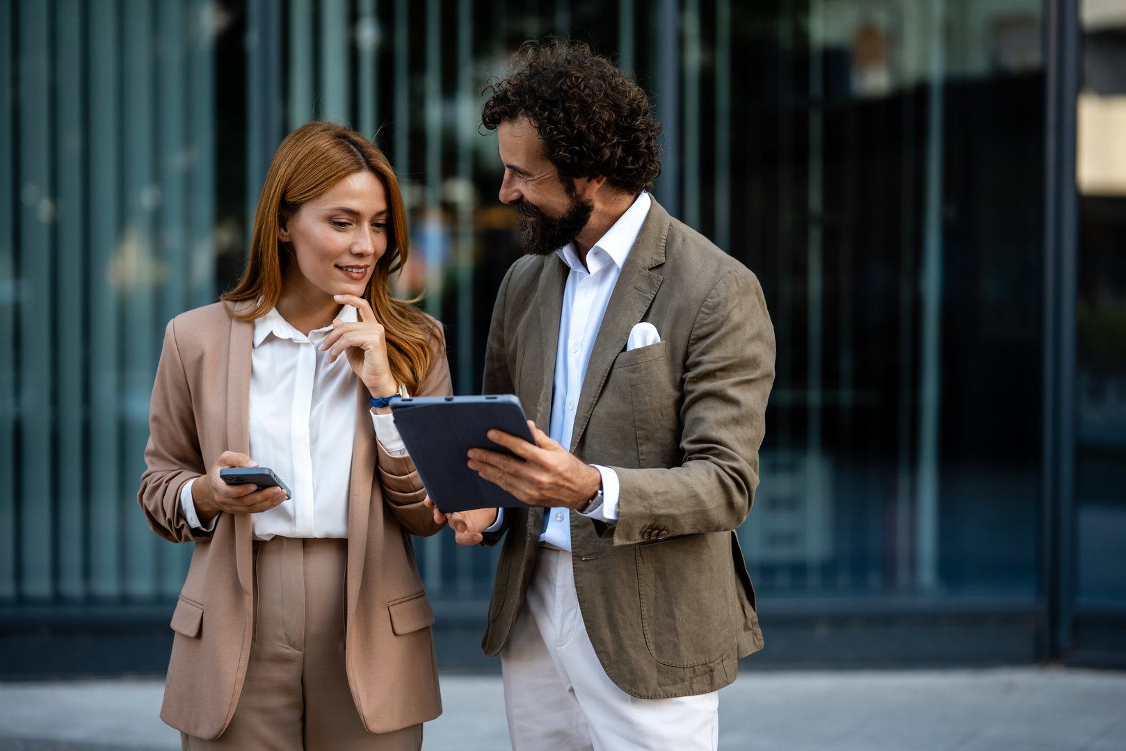 Business partners reviewing project details using a digital tablet and smartphone in front of a modern office building