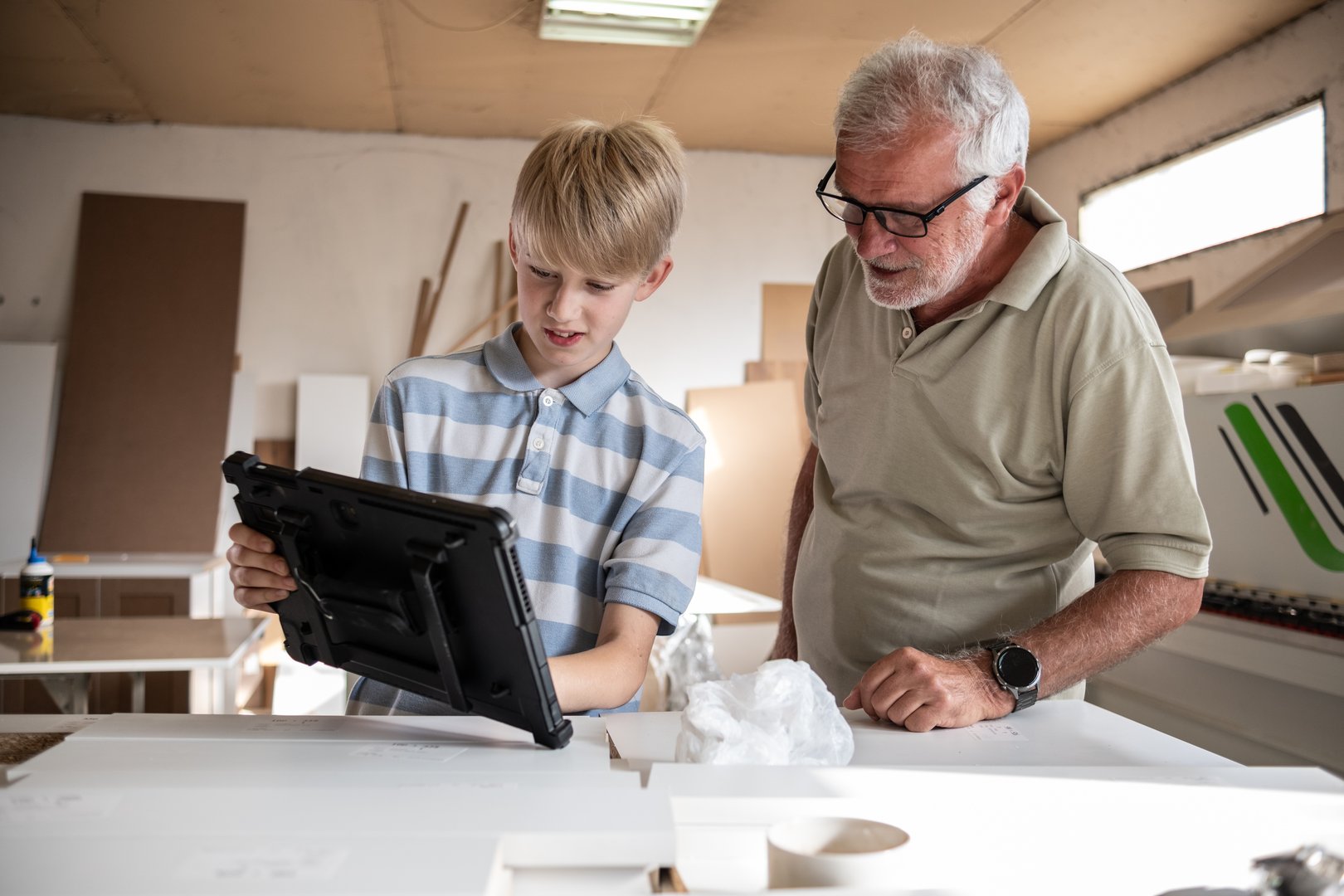 Grandson teaching his grandfather carpenter how to use a tablet to improve their work. The grandson shares his tech knowledge, helping the grandfather integrate modern tools into their craft.