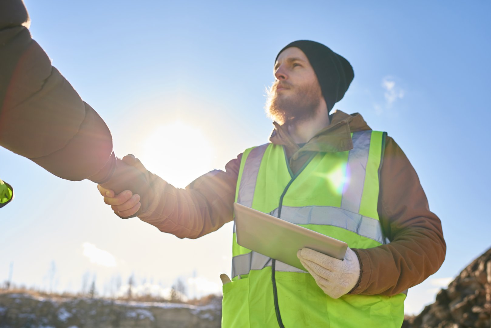 Low angle portrait of bearded industrial worker wearing reflective jacket shaking with partner hands outdoors against cold blue sky
