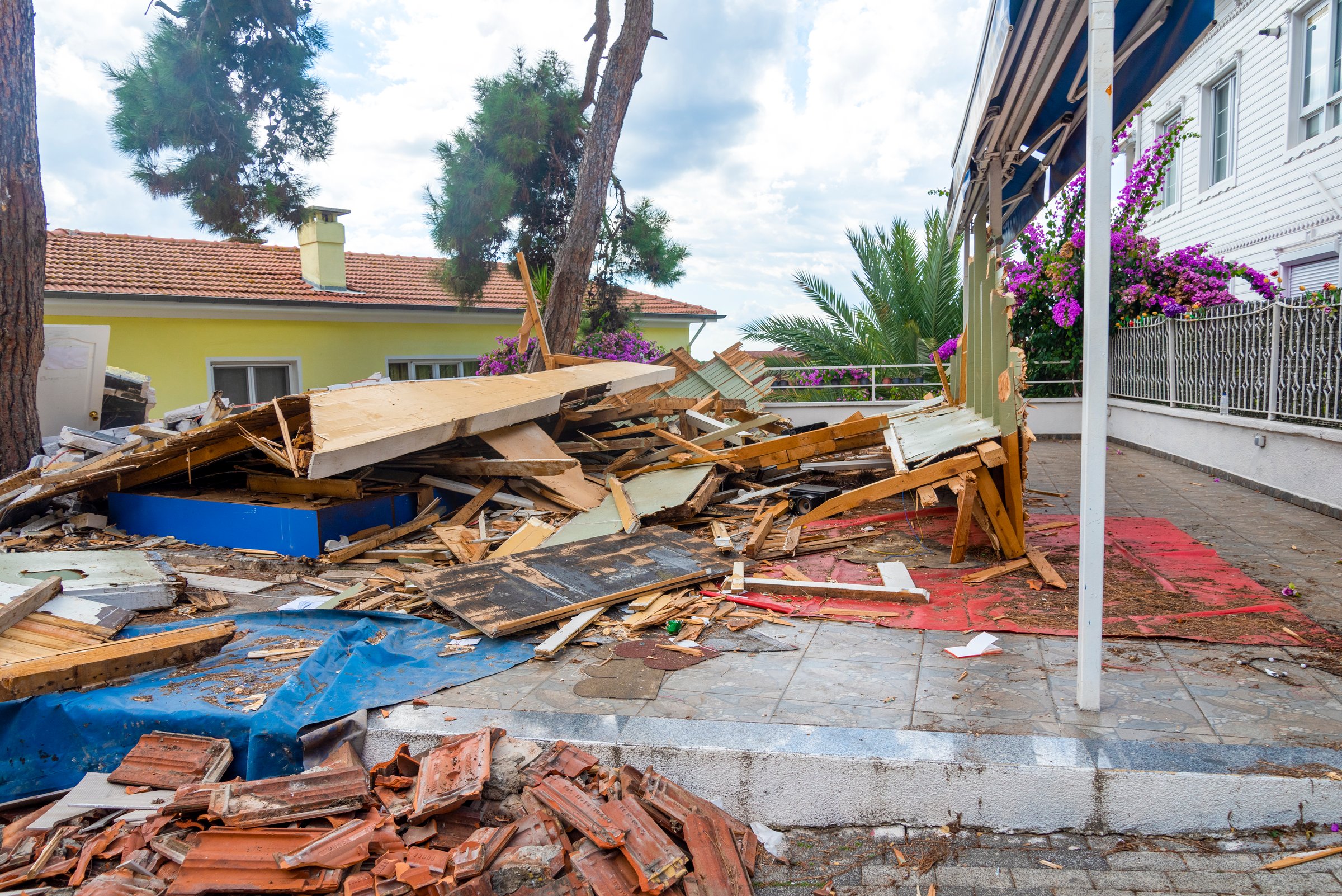 A significant amount of debris lies scattered on the ground where a structure once stood. Surrounding plants and homes are visible, showing signs of recent construction activity.