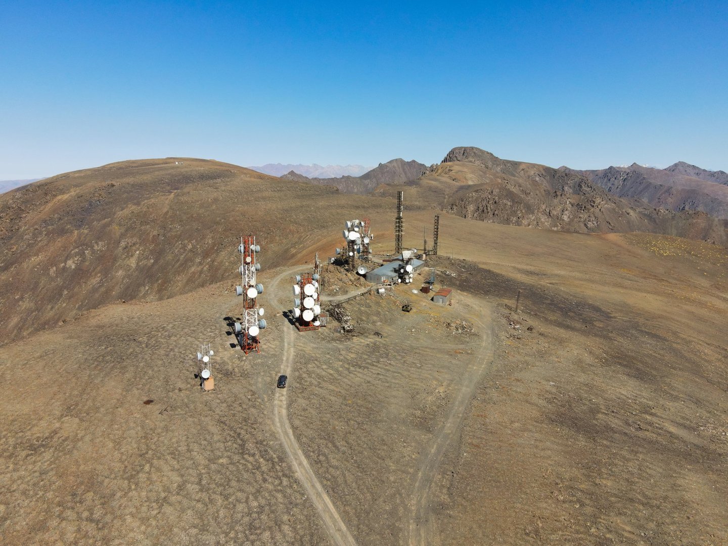 An aerial view showing multiple communications towers on a mountaintop, with a small black vehicle visible on the dirt tracks leading to the facility