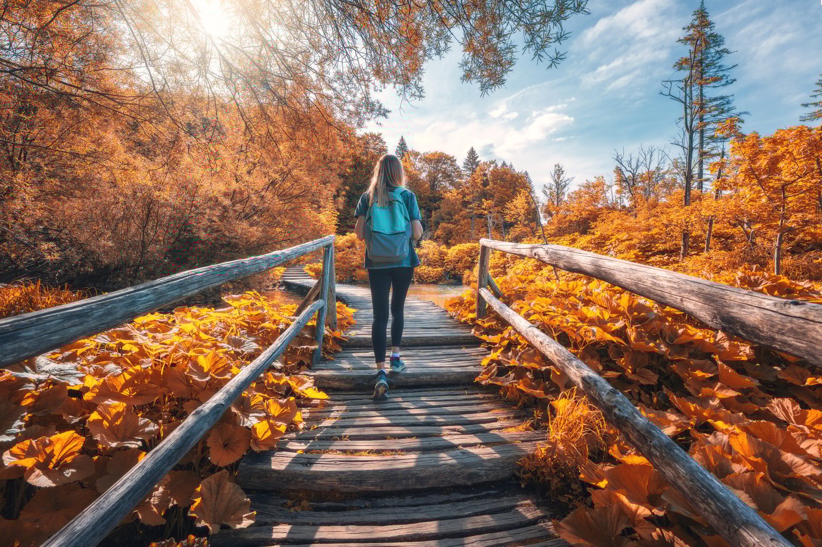Young woman with backpack walking along a wooden path near stunning waterfalls, orange trees in Plitvice Lakes national park, Croatia at sunset in autumn. Sporty girl in forest in fall. Hiking, travel