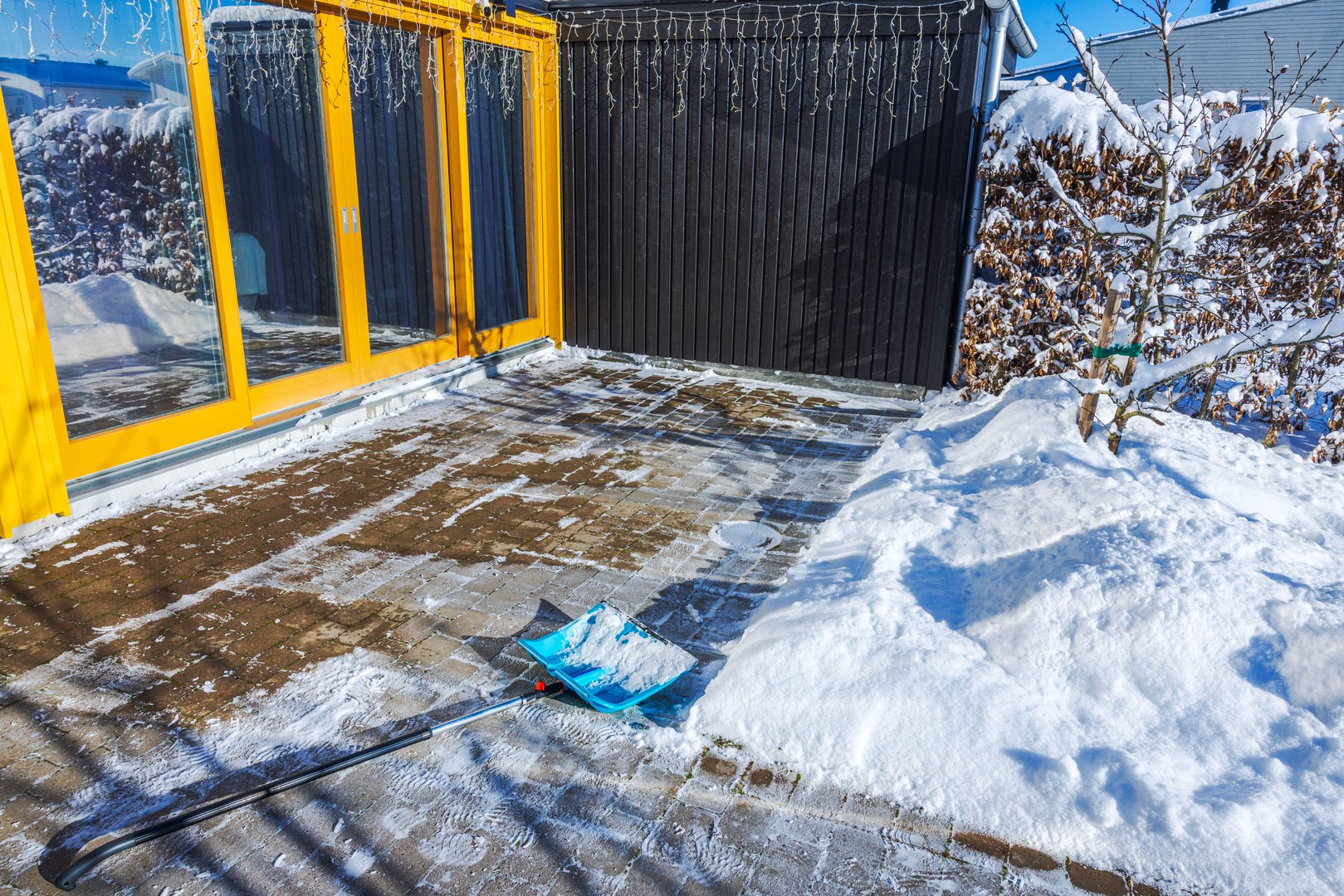 Blue snow shovel lying on cleared brick patio near panoramic windows of villa on sunny winter day. Sweden.