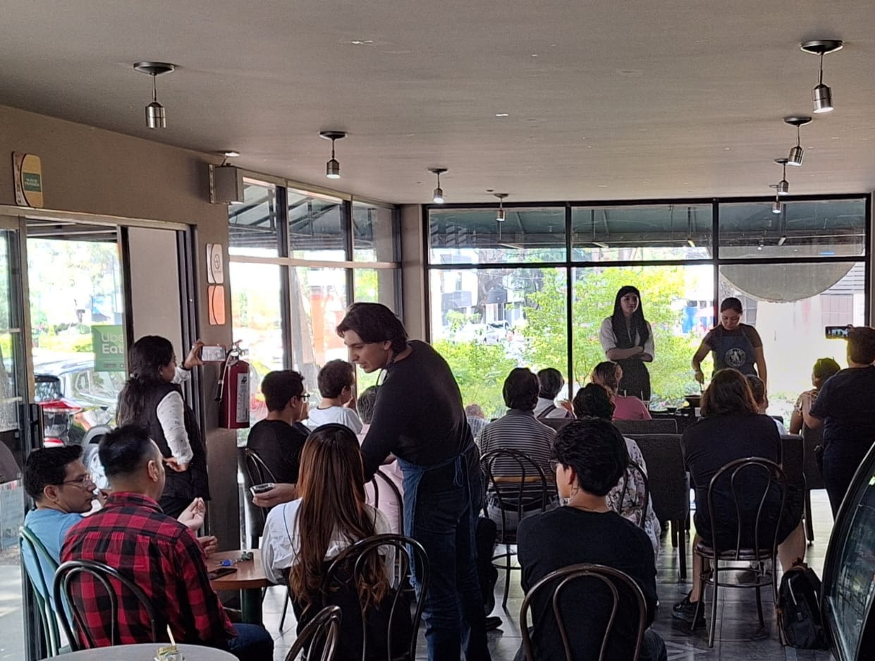 A group of people seated indoors at a community event with two presenters speaking at the front.